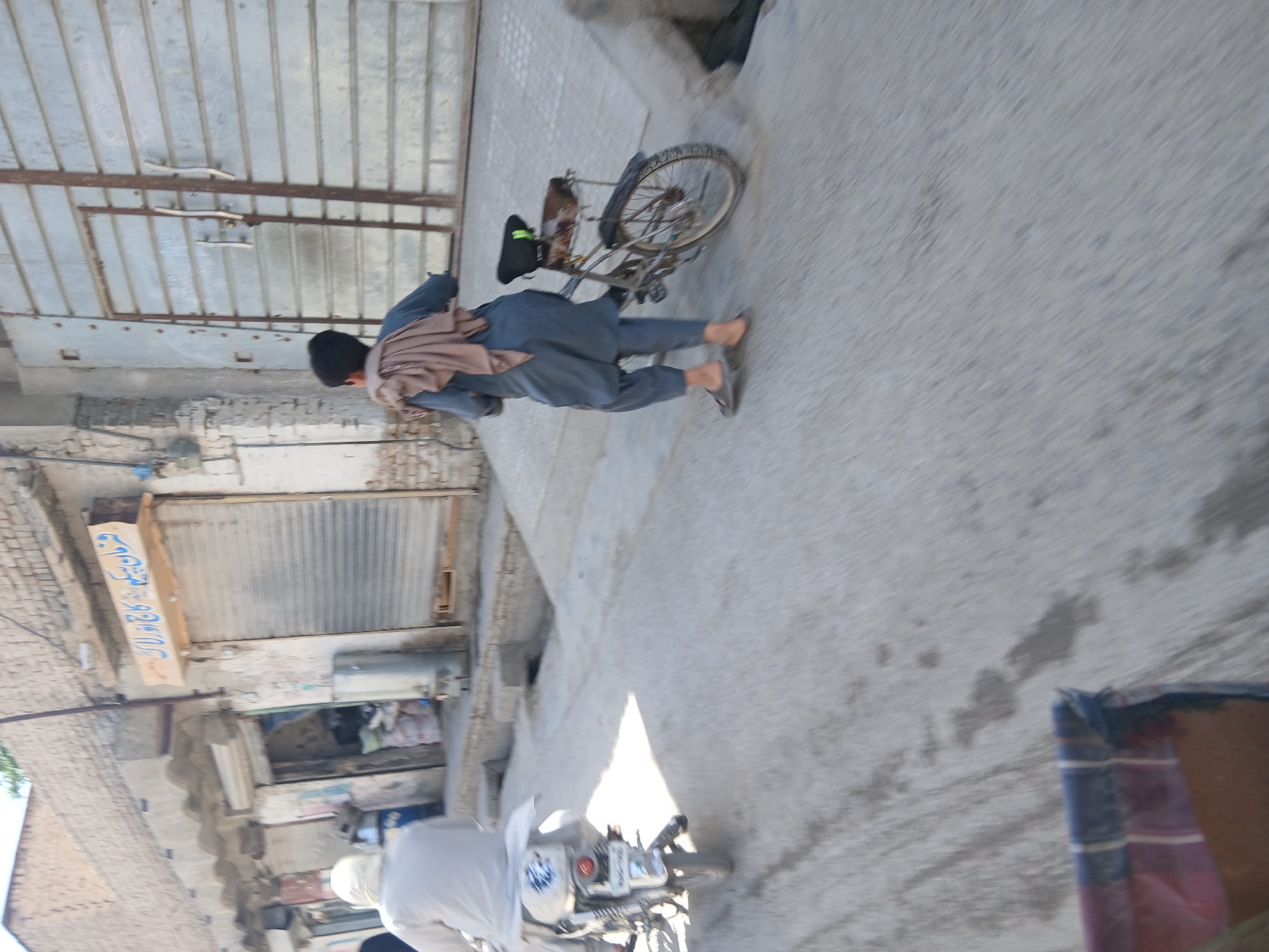 A slightly blurry photo shows a man walking with a bicycle down a city street in Quetta City Tehsil, Pakistan. The man is wearing blue clothes, a brown shawl and black cap with sandals on his feet. He is wheeling a bicycle. In the foreground, another man riding a motorcycle is visible. The shops on the side of the road are shuttered with some text written on the side of one shop.