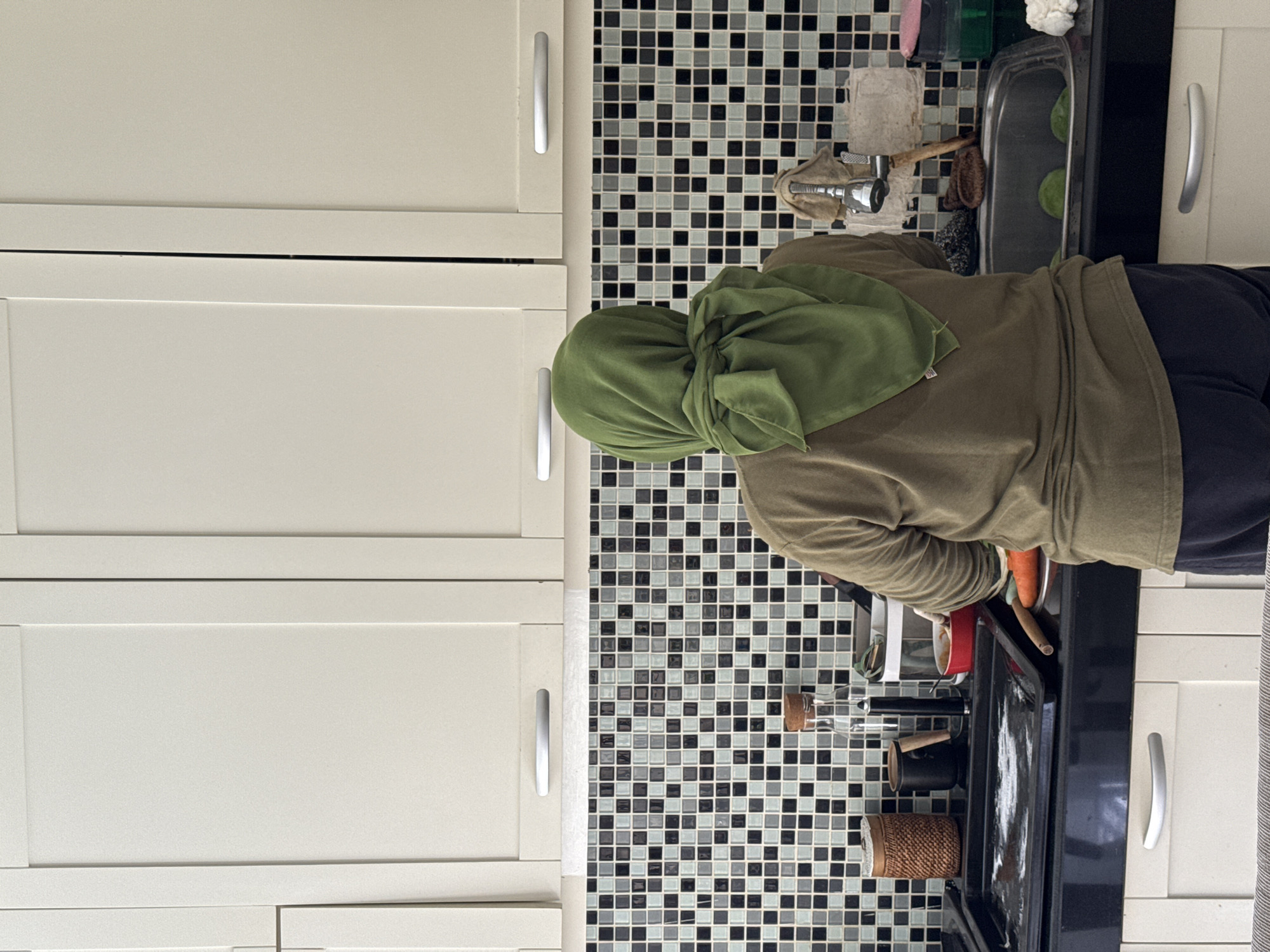 A person wearing a green headscarf and an olive-green long-sleeved shirt stands in a kitchen, seemingly engaged in cooking or preparing food. The kitchen features white cabinets with silver handles and a backsplash composed of small, square black, white, and gray tiles. A sink with a silver faucet and various cleaning supplies sits to the right. The person is facing the stove, where cookware and utensils are present. The location is likely indoors, suggesting a private residence in the Kuta Selatan, Indonesia area.