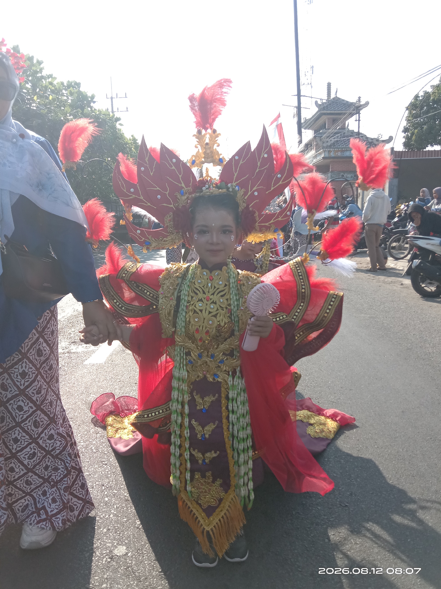 The image depicts a young girl adorned in a vibrant, traditional costume, possibly for a parade or cultural event, standing on a paved street in Gudo, Indonesia. The costume is predominantly red and gold, featuring intricate embellishments, feathered headwear, and a long, flowing skirt. She is holding a small, pink handheld fan. To her left, an adult's arm is visible, with their hands clasped together, suggesting they are walking with the child. In the background, a traditional Indonesian building with a tiled roof can be seen, along with other people, some on foot and others near motorcycles. The scene appears to be outdoors on a sunny day, with shadows cast on the street. A date stamp in the lower right corner reads "2026.08.12 08:07".