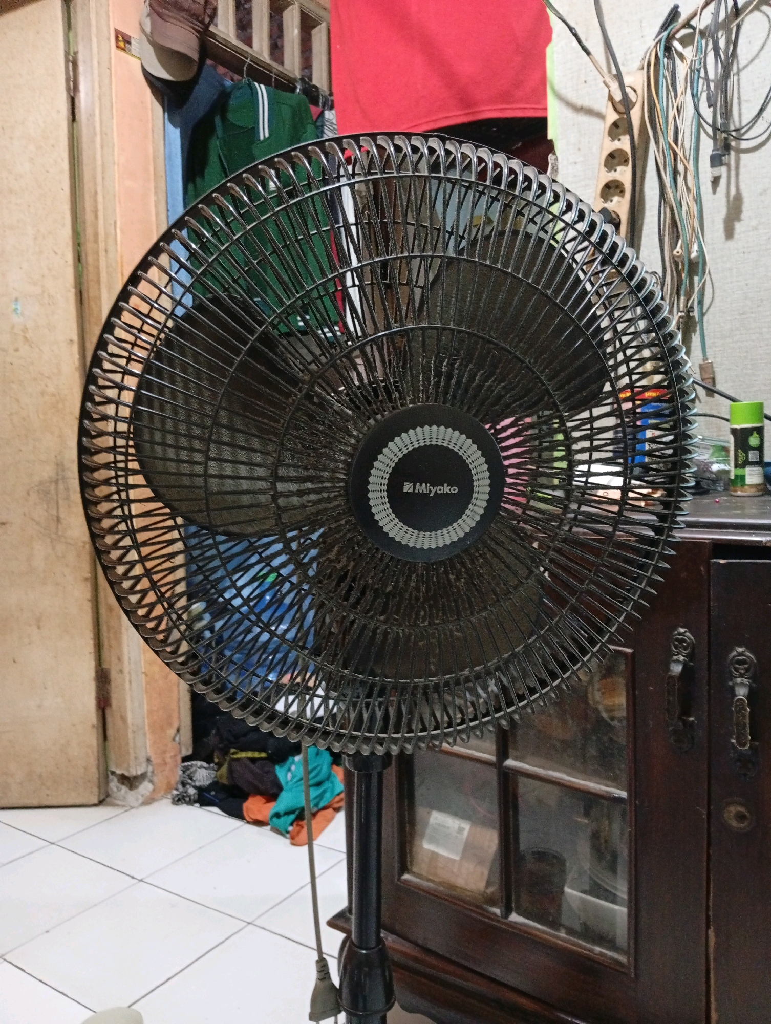 This is a medium shot, eye-level, still photograph of a black Miyako brand electric fan in the foreground, positioned in a room. The fan is oriented to the right, and its blades are visible through the black grate. The fan has a circular emblem with the Miyako logo in the center. Behind the fan, on the left side of the image, a doorframe is visible. To the right of the fan, a dark wooden cabinet with two drawers is present. The cabinet has ornate handles and a glass window on the top drawer, through which various objects are visible, including what appears to be photographs and a cylindrical object. Above the cabinet, there is a shelf with various items, including a green spray can and other indistinct objects. The wall behind the fan and cabinet is textured. In the background, further to the left, a pile of colorful clothing is on the floor. To the upper left of the fan, clothing is hanging on a rack. The floor is tiled in a white, square pattern. The scene appears to be indoors in a residential setting in Caringin, Indonesia, likely a bedroom or living area, characterized by clutter and everyday household items.