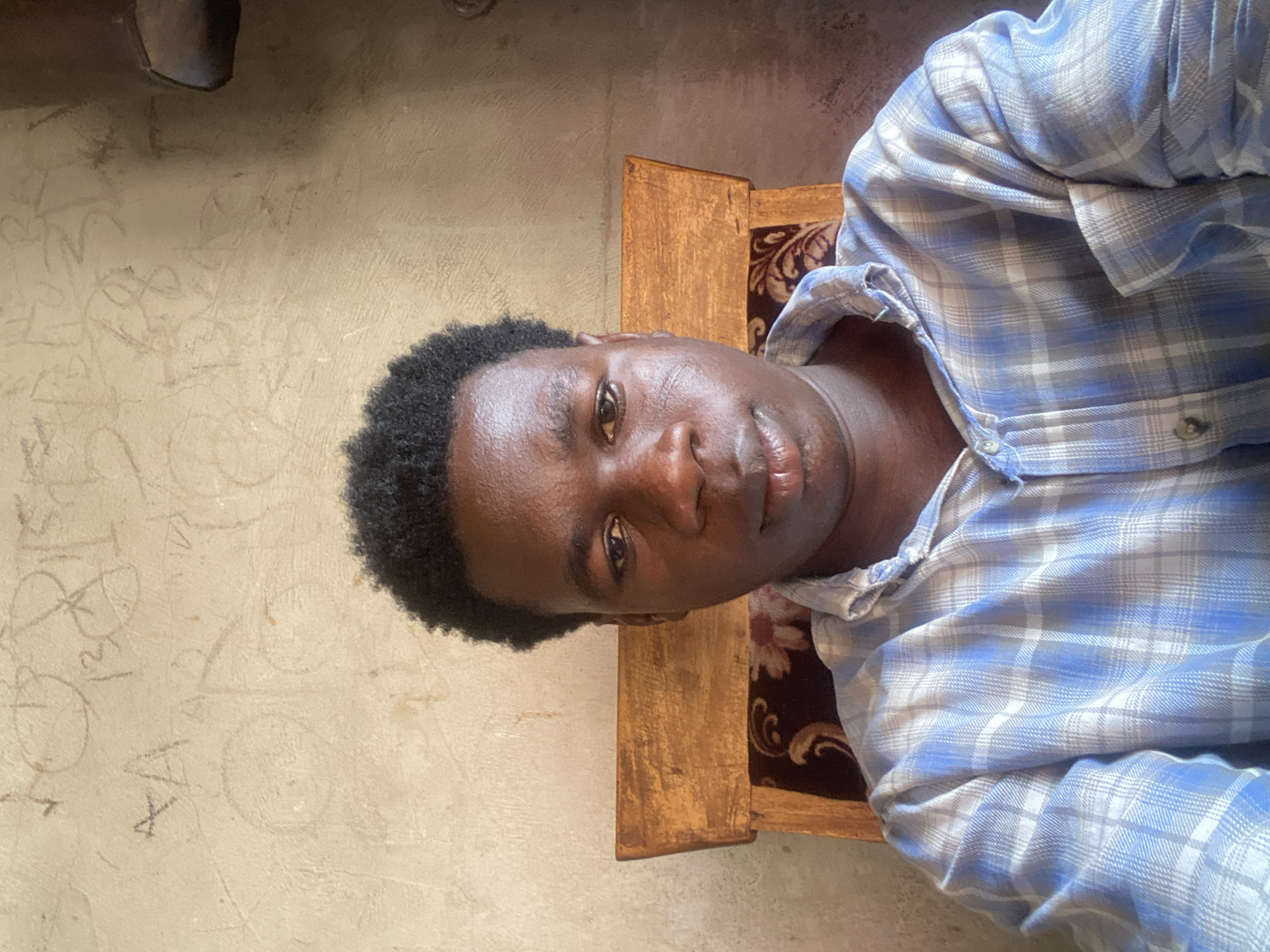 A medium indoor shot shows a dark-skinned person with a short, curly haircut, wearing a blue and white plaid shirt. The individual is sitting in a wooden chair with a dark patterned fabric backrest. The background features a light-colored wall with faint, scribbled markings. The location is presumed to be in Mararaba, Nigeria.