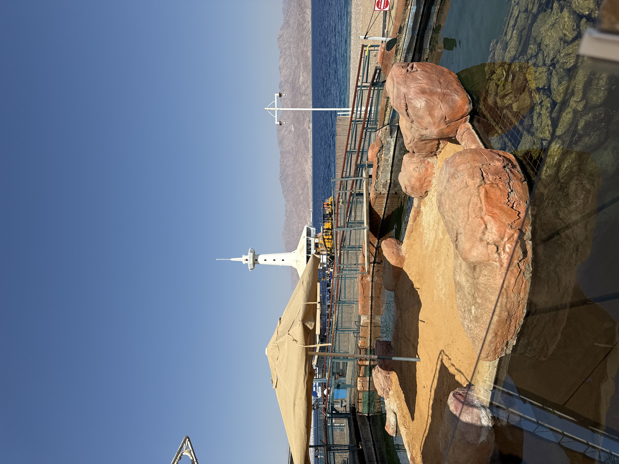 The image depicts an outdoor exhibit at the Coral World Underwater Observatory in Eilat, Israel. In the foreground, a sandy enclosure with large, reddish-brown rocks sits beside a clear water pool. An umbrella shades a portion of the sand. A wooden walkway with a metal railing spans across the exhibit, leading towards the background.

Beyond the exhibit, the Red Sea is visible, with a sandy beach in the distance and a mountain range forming the horizon under a clear blue sky. A white tower, characteristic of the observatory, stands prominently near the water's edge. Several boats and yellow structures are also present on the water. The overall scene suggests a sunny day at a tourist attraction focused on marine life.