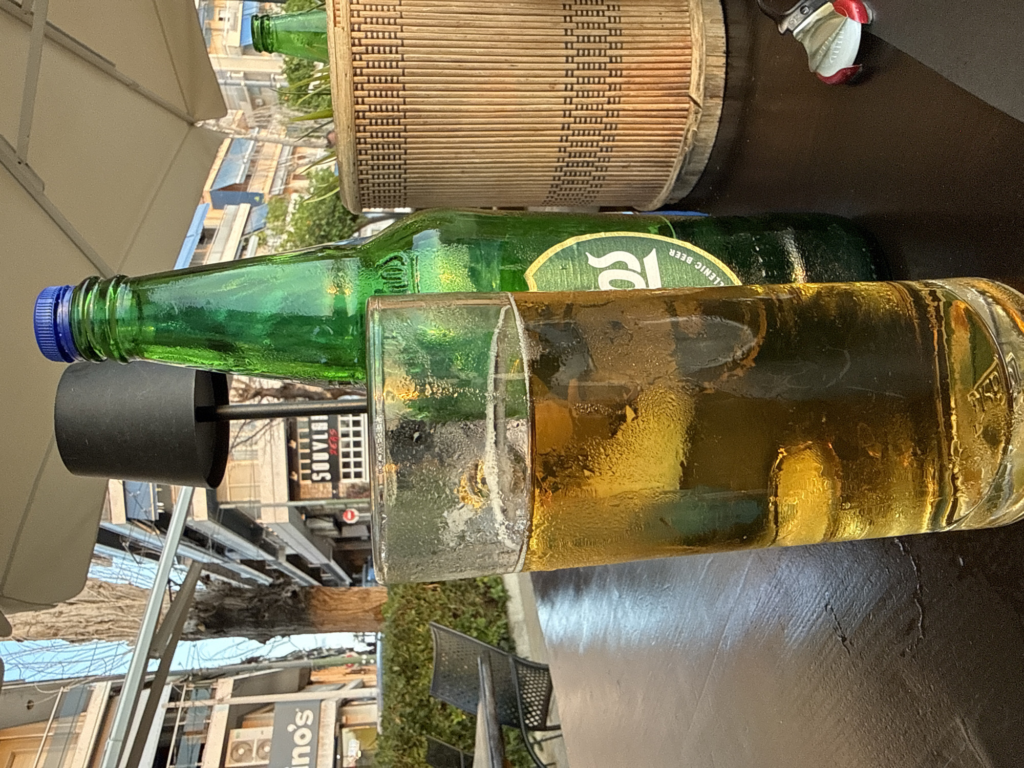A green beer bottle with a blue cap lies next to a glass of beer on a table. A wicker planter and an outdoor chair are nearby. The scene is likely at an outdoor seating area in Piraeus, Greece, suggested by signs visible in the background including "Souvlaki", "Islandos" and "ino's".