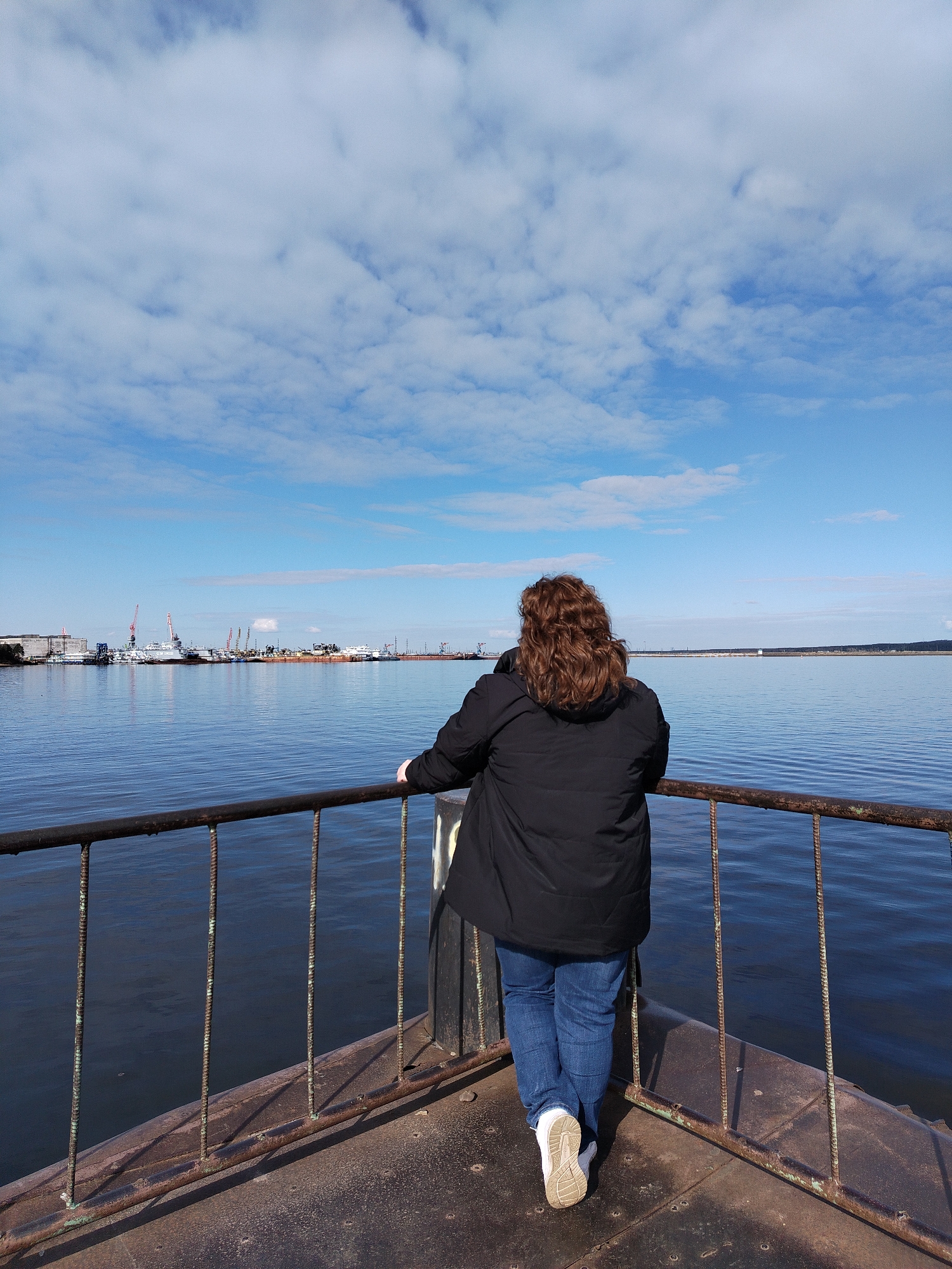 A person stands on a rusted, metal pier, looking out over a body of water under a cloudy sky. The water stretches horizontally to the horizon. On the distant shore, there are port facilities including cranes. The person is facing away from the camera, wearing a black jacket, blue jeans, and white shoes. The rusty metal pier features a simple railing. This image appears to be taken in the city of Chaikovsky, Russia.