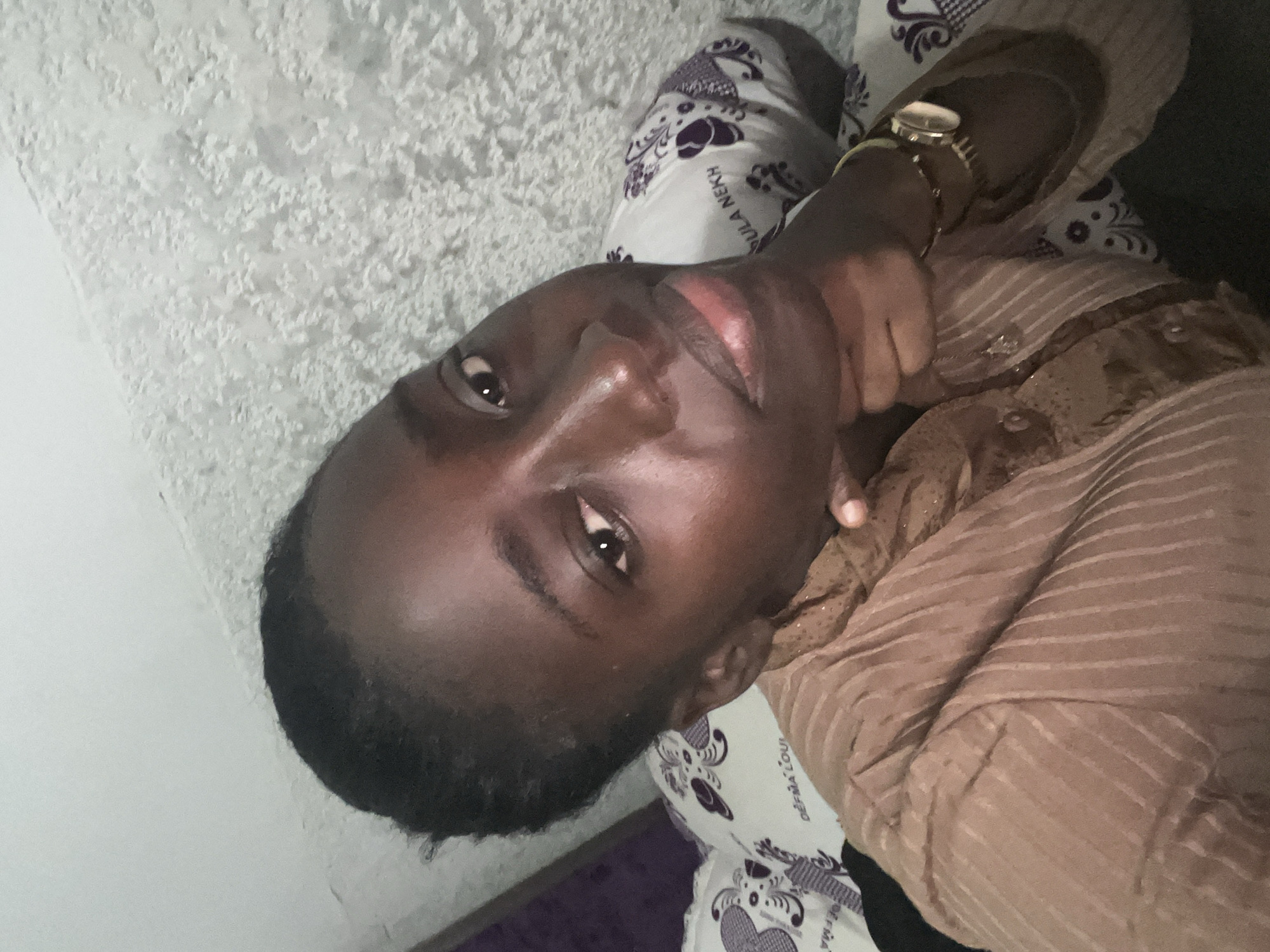 The image shows a dark-skinned person with short, dark hair lying on a patterned fabric. The person is wearing a brown, striped shirt and a watch on their left wrist. The patterned fabric appears to be a pillow or bedding with purple designs on a white background. The background includes a textured wall that looks like it could be stucco or a similar material. There is no indication of a specific location in Abuja, Nigeria.