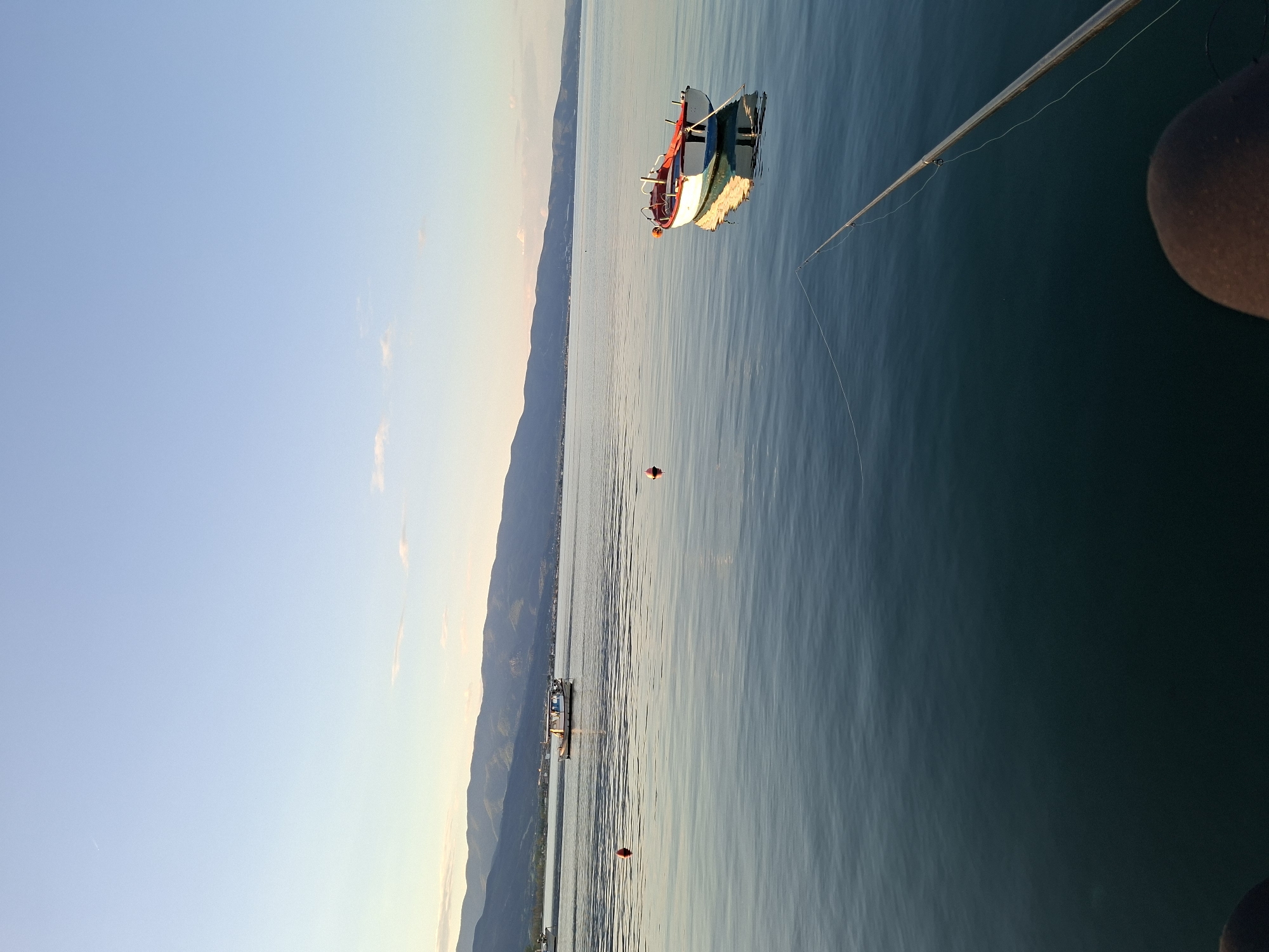 The image, oriented horizontally, captures a tranquil water scene during either sunrise or sunset. In the immediate foreground, from the bottom right corner, a fishing rod extends over the water, with its line clearly visible and cast. A dark, rounded object, likely part of a person's shoulder or a boat component, is partially visible in the extreme bottom right, suggesting someone is fishing.
The calm, blue-green water dominates the mid-ground, featuring gentle ripples that reflect the soft light. A small, colorful boat, painted white with red and blue trim, floats peacefully on the water, its reflection subtly visible beneath it. Further into the water, two small red buoys are scattered, with one appearing closer to the boat.
In the distance, a long mountain range or series of hills stretches across the horizon, bathed in warm, soft light. The sky above is a clear, light blue, gradually transitioning to a lighter, warmer hue near the distant land. The overall atmosphere is one of serenity and peace. There is no visible text in the image.