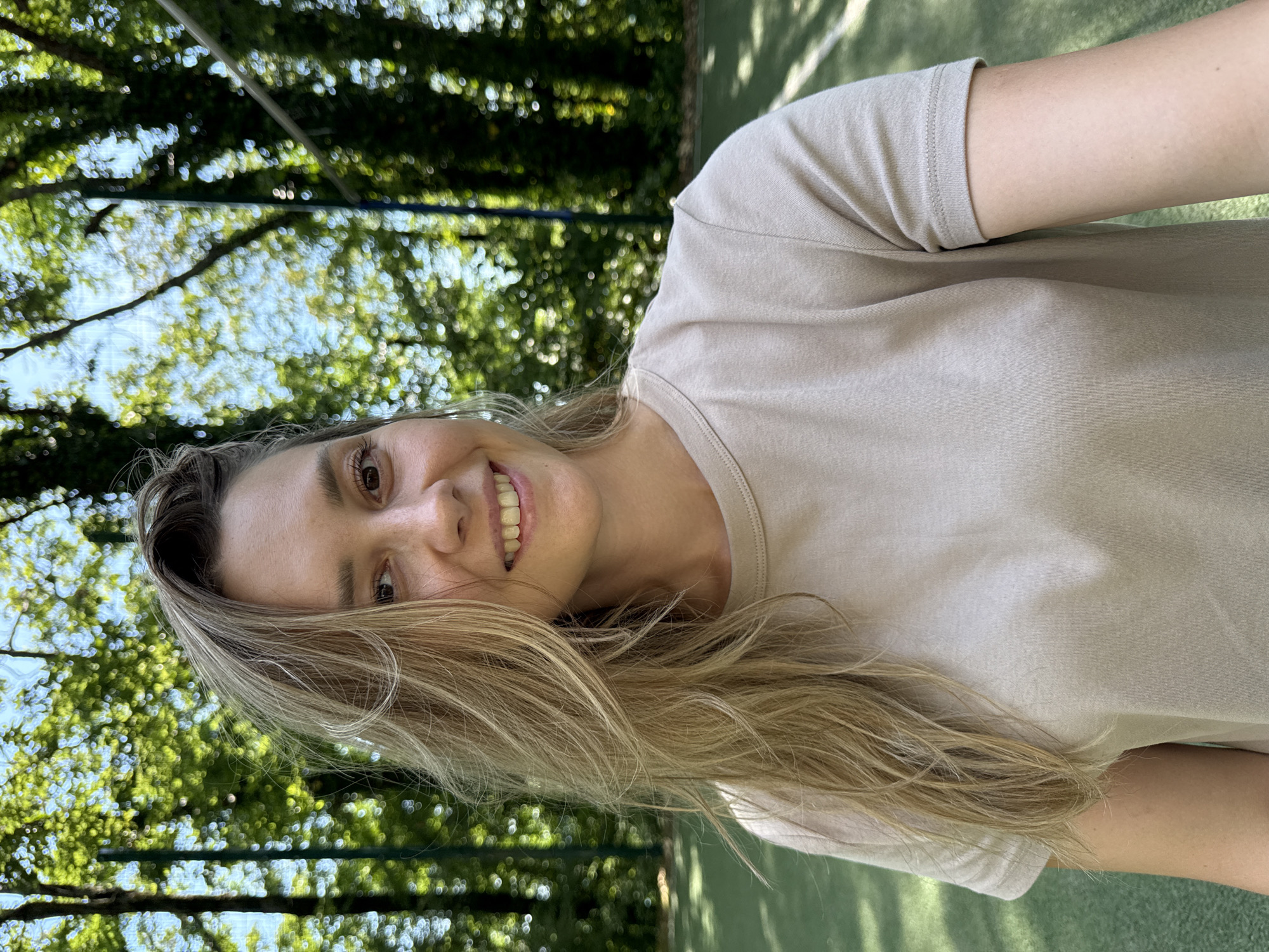 A woman with long, blonde, wavy hair is pictured from the chest up, outdoors, in a selfie. She is smiling and looking directly at the camera. She is wearing a light brown, short-sleeved t-shirt. Behind her, the background consists of lush green trees and a bright blue sky, suggesting a sunny day. A portion of a green court, possibly a tennis court, is visible in the lower part of the image, along with a blurred net or fence. The overall setting appears to be a park or recreational area.