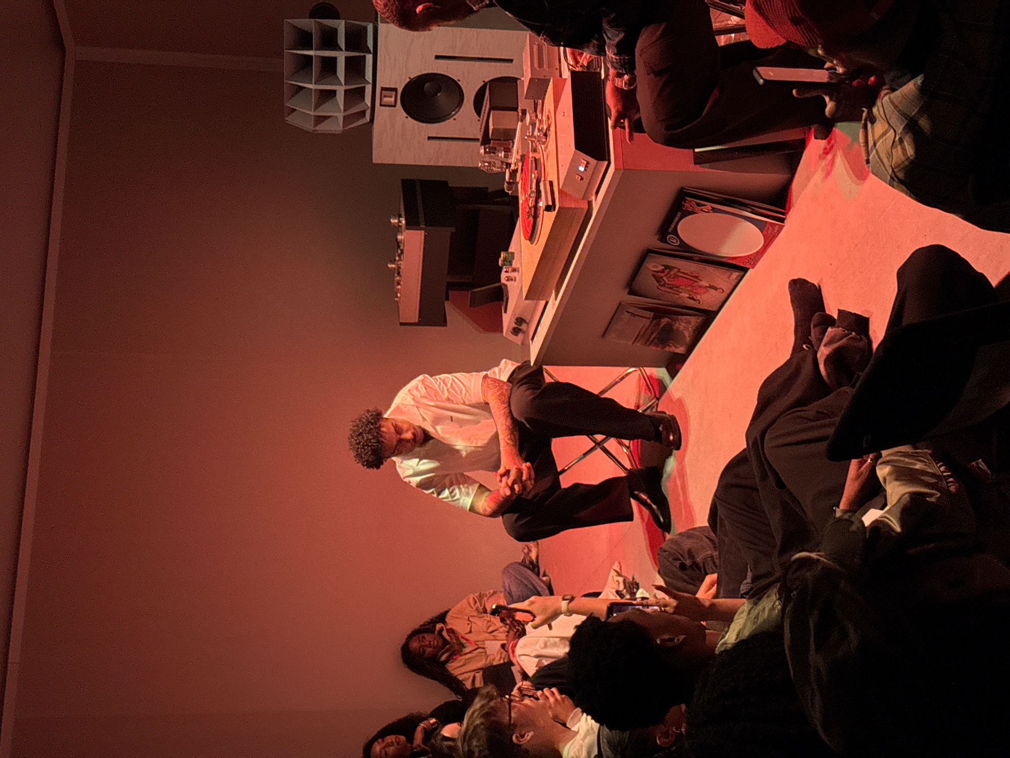 The image shows a gathering of people in a room illuminated by red lighting, seemingly focused on a man sitting on a metal chair near a DJ setup. The man is wearing a white shirt and black pants, with visible tattoos on his arms. He appears to be either speaking or pausing in contemplation. A group of people is situated on the floor, seemingly watching or listening intently. A DJ setup featuring a turntable, mixer, and speakers is present, along with album covers on the desk below. Based on the distinctive speaker cabinet and the general style of the event, the location is likely a listening session or a small performance related to music, specifically vinyl culture. Without more evidence, it is not possible to definitively confirm its location within London.