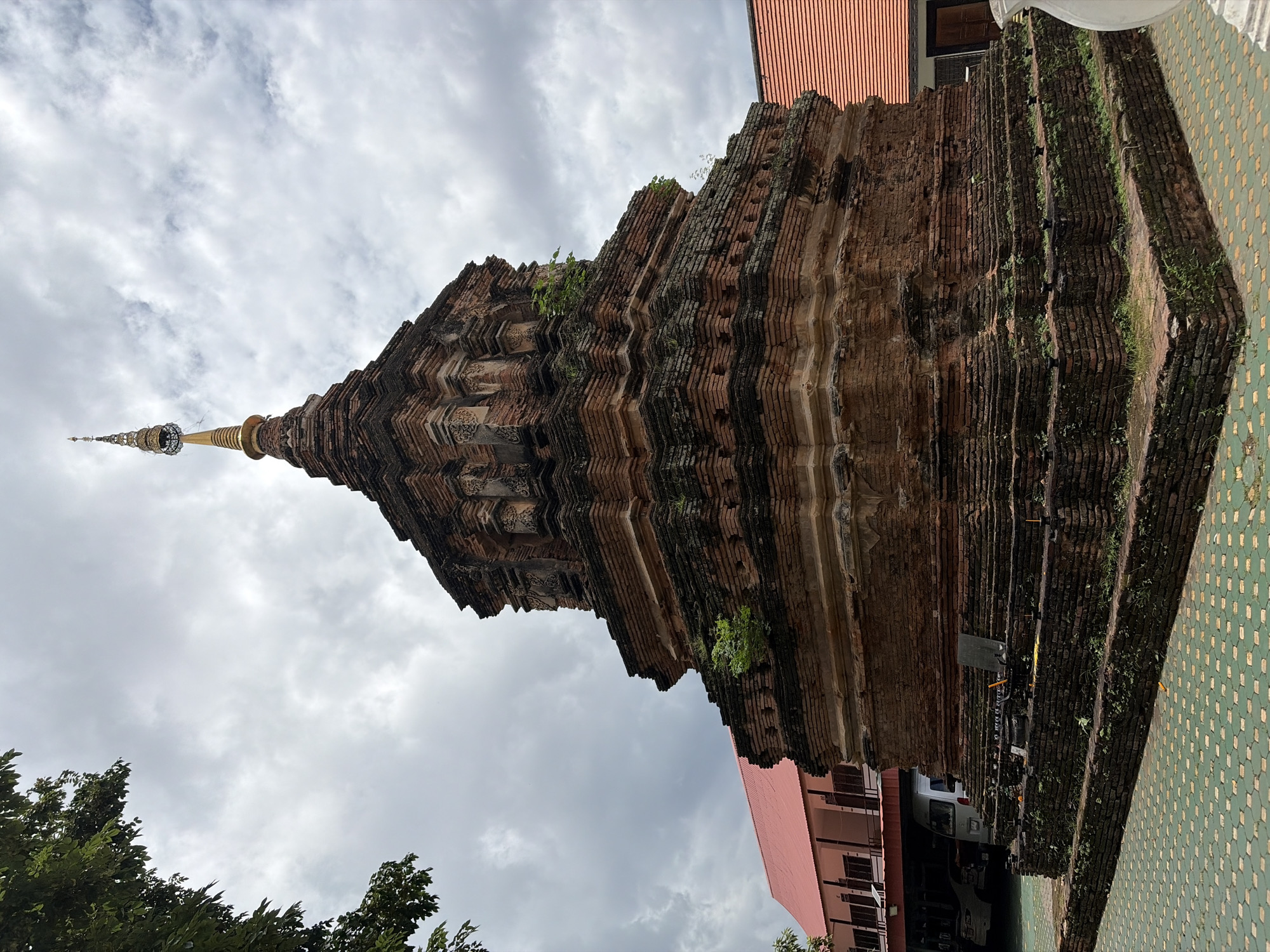 A Buddhist stupa, constructed from weathered red bricks, stands prominently in the foreground against a cloudy sky. The stupa features multiple tiered levels, with intricate carvings and remnants of moss and small plants growing on its surfaces. A golden spire adorns the apex of the stupa. To the left of the stupa, a building with a red roof and light pink walls is partially visible, with parked vehicles, including a white van, beneath its covered area. To the right, another building with an orange tiled roof is visible. The ground surrounding the stupa is paved with green and yellow hexagonal tiles. The overall scene suggests a temple or historical site within Chiang Mai City Municipality, Thailand.