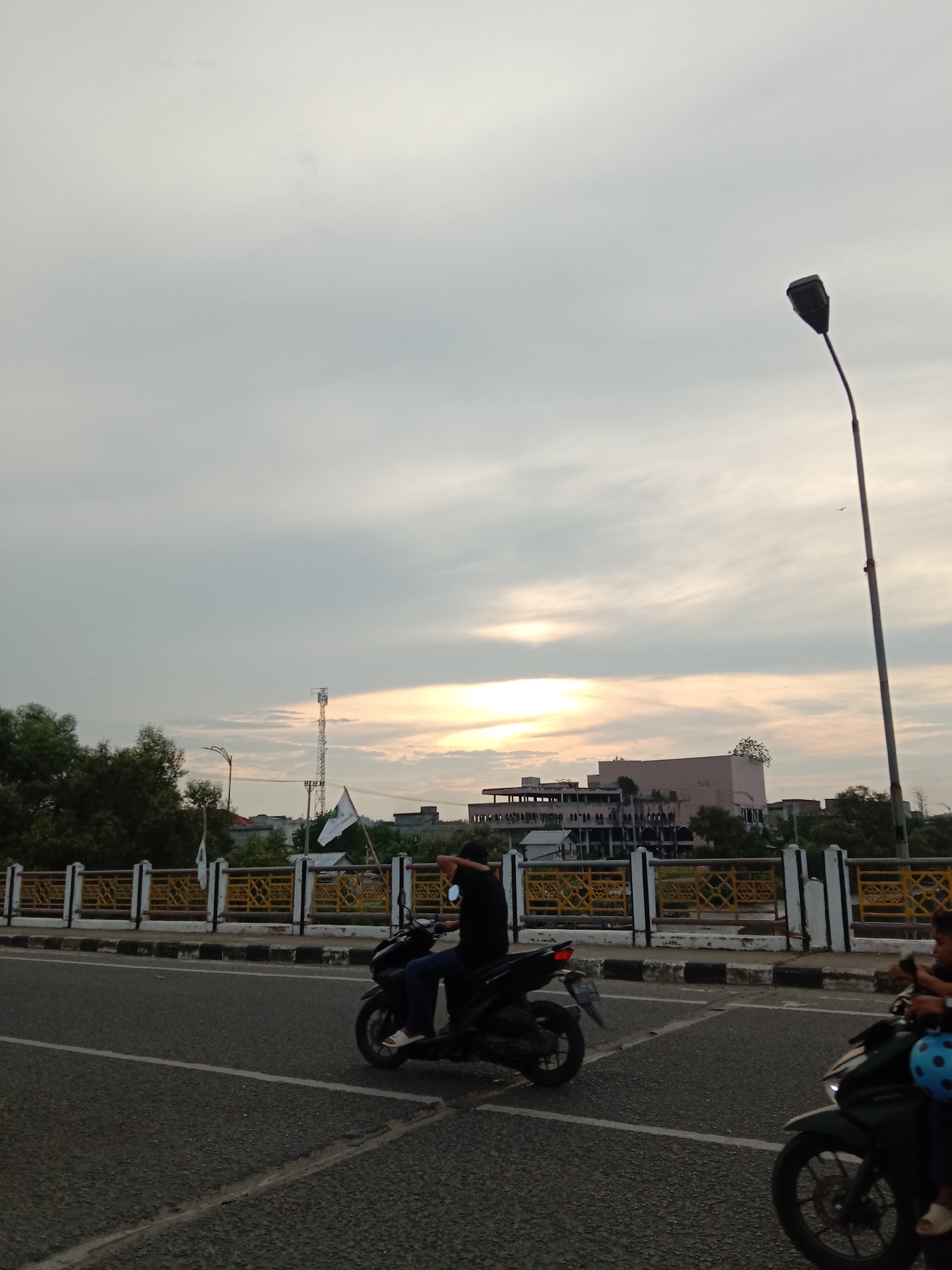 The image shows a street scene in Lhokseumawe, Indonesia. Two people are riding motorcycles. A tall street light stands on the right side of the frame. In the background are trees, a building under construction, a cell tower, and a bridge with a yellow railing. The sky is cloudy. A person on a motorcycle is holding a white flag.