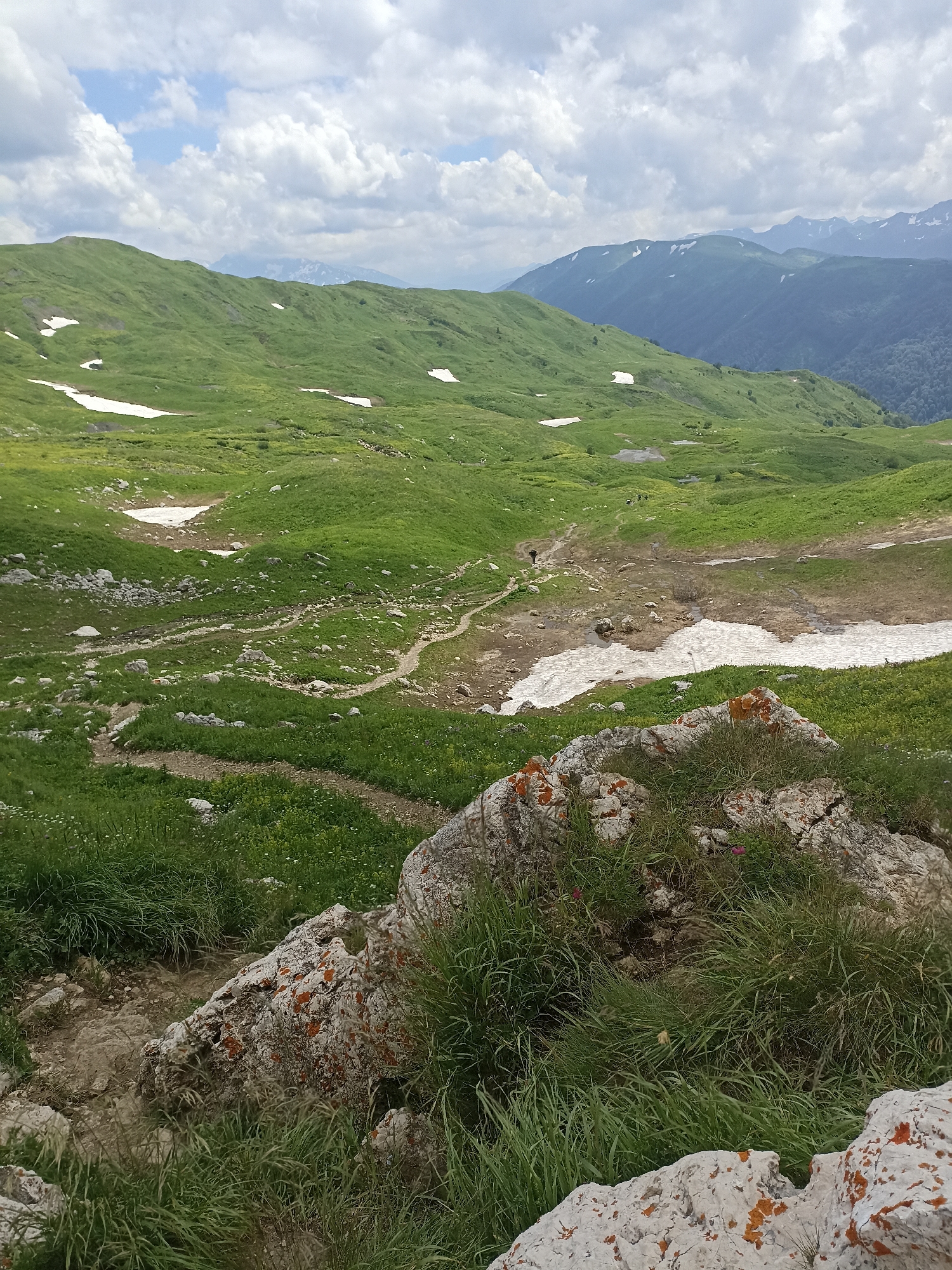 The image depicts a vast, mountainous landscape with rolling green hills and patches of snow. The sky is filled with white, fluffy clouds against a light blue backdrop. In the foreground, large rocks covered in orange lichen and green grass are visible. A dirt path winds through the terrain, leading towards a shallow body of water or wet ground. In the distance, several figures, appearing small due to the distance, are walking along the path. Further in the background, more mountain ranges are visible, some with snow-capped peaks. The overall environment suggests a natural, outdoor setting, likely a hiking trail in a mountainous region. The location provided, Chigursana, Russia, indicates a high-altitude area with mountainous terrain.