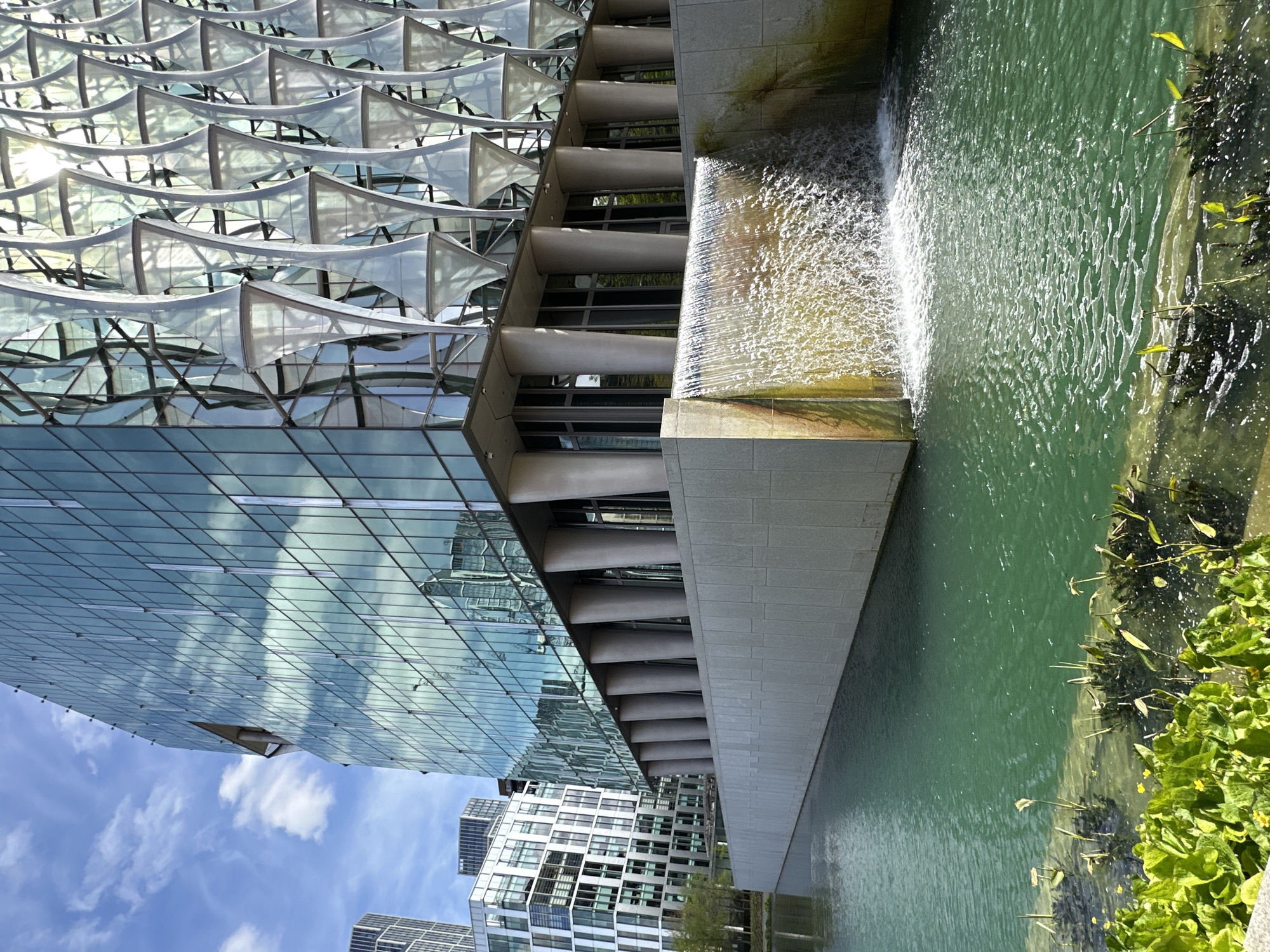 The image depicts a modern architectural scene in London, United Kingdom, specifically focusing on a building with a distinctive facade. The facade features a pattern of white, sail-like structures over glass panels. Adjacent to the building, there is a water feature with a cascading waterfall flowing into a pond. Green vegetation grows along the water's edge. The sky is visible in the background, featuring blue hues and scattered clouds.