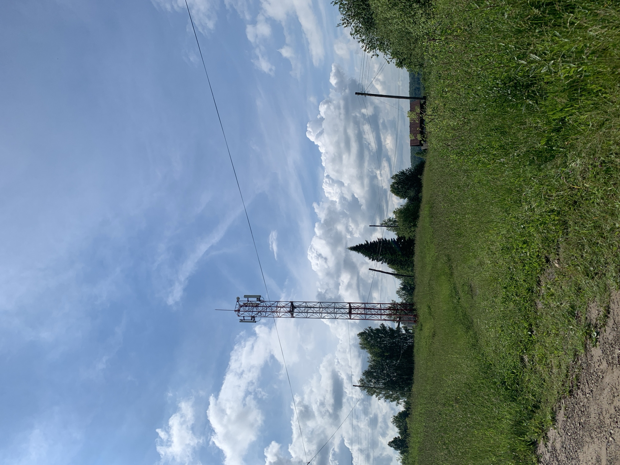 The image depicts a cellular tower standing tall in a grassy field under a partly cloudy sky. The tower, predominantly red and grey metal lattice, is equipped with multiple antennas. A dirt path leads towards the tower through the lush green grass. In the background, a line of trees separates the field from a small cluster of buildings, one of which has a rusty brown roof. Power lines crisscross the scene, supported by wooden poles. The overall environment suggests a rural or semi-rural setting in the city of Barsai, Russia. No people are visible in the frame.