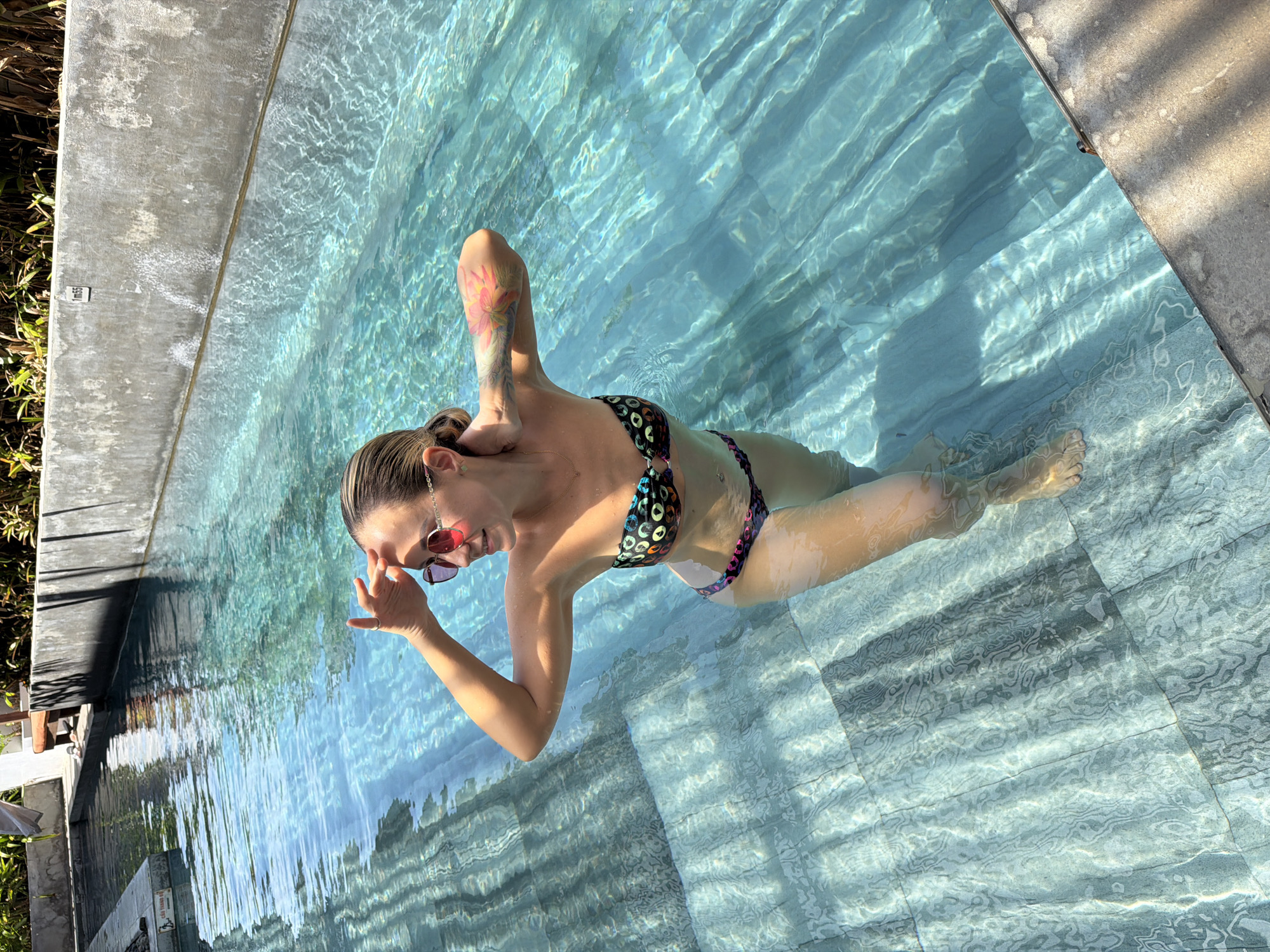 The image shows a woman in a swimming pool. She is wearing a bikini and sunglasses. Her body is partially submerged in the clear blue water, which is illuminated by sunlight, creating rippling patterns on the pool's tiled bottom. She has tattoos on her arm and is touching her face. The pool area appears to be outdoors, with concrete edges and some greenery visible. The lighting suggests it is daytime. The scene conveys a sense of relaxation and enjoyment. A sign with text is visible on the concrete edge of the pool, but the text is too small to be legible.