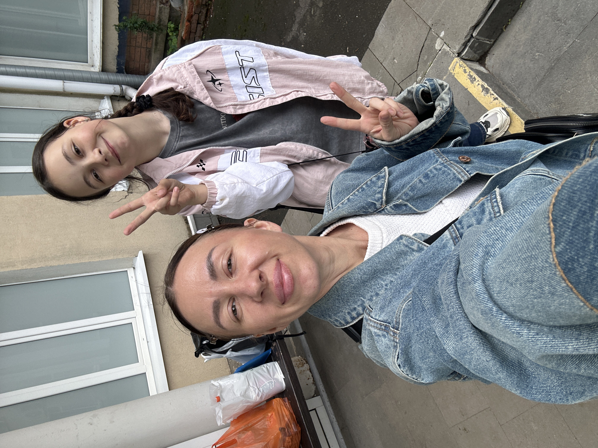 The image features two smiling females of different ages posing outdoors, likely for a selfie, with the image rotated 90 degrees clockwise. Both are making a peace sign (V-sign) with one hand.
In the foreground, an older woman with dark hair pulled back is wearing a light-colored, possibly white, knit sweater under a blue denim jacket. She has a pleasant, happy expression.
Behind her, a younger girl with long dark hair, part of which is braided and secured with a dark scrunchie, also smiles at the camera. She wears a grey t-shirt under a pink and white windbreaker-style jacket. The word "FIRST" with a star logo is visible on her jacket, and "OUTDOOR" can be partially seen on her t-shirt. Her blue jeans and a white sneaker are also visible.
The setting is an outdoor urban or suburban environment, possibly an alley or sidewalk next to a building. The background includes a light-colored building facade, a white-framed window, and sections of brickwork with some green foliage. The ground is a grey paved surface with a distinctive yellow painted line. Several plastic bags (white, orange, blue) and a black bag are on the ground near the older woman. The lighting is even, suggesting an overcast daytime.