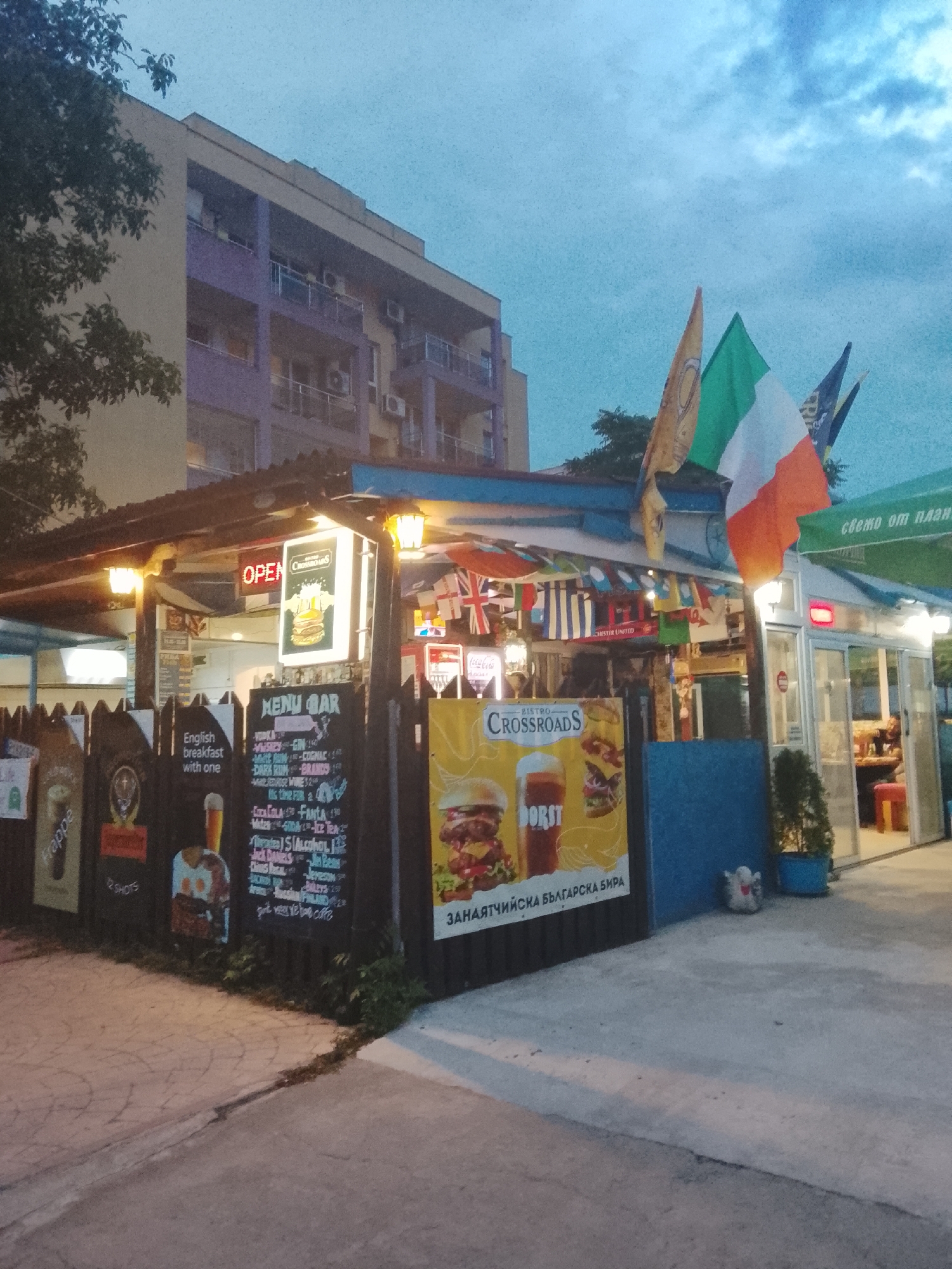 This image captures a street-level view of a bar or cafe, likely located in Nesebar, Bulgaria, given the presence of a sign in Bulgarian text that reads "ЗАНЯТИЧНА БЪЛГАРСКА БИРА" which translates to "Bulgarian Beer". The establishment appears to be open, indicated by a glowing red "OPEN" sign. The facade features a covered outdoor seating area with a rustic, wooden fence-like structure displaying various menus and advertisements.
Dominating the scene are several flags, including the Irish flag and potentially the flag of England, hung from the roofline. These flags, along with other smaller international flags adorning the structure, suggest a place with an international theme or clientele. One prominent advertisement displays an image of a burger and the text "BISTRO CROSSROADS", indicating the name of the establishment.
The time of day appears to be twilight, with a dusky sky and artificial lighting illuminating the area. In the background, a multi-story building, possibly an apartment complex or hotel, is visible, characterized by its purple and beige facade. The ground in front of the establishment is paved with concrete. The overall impression is that of a casual, open-air dining and drinking venue.