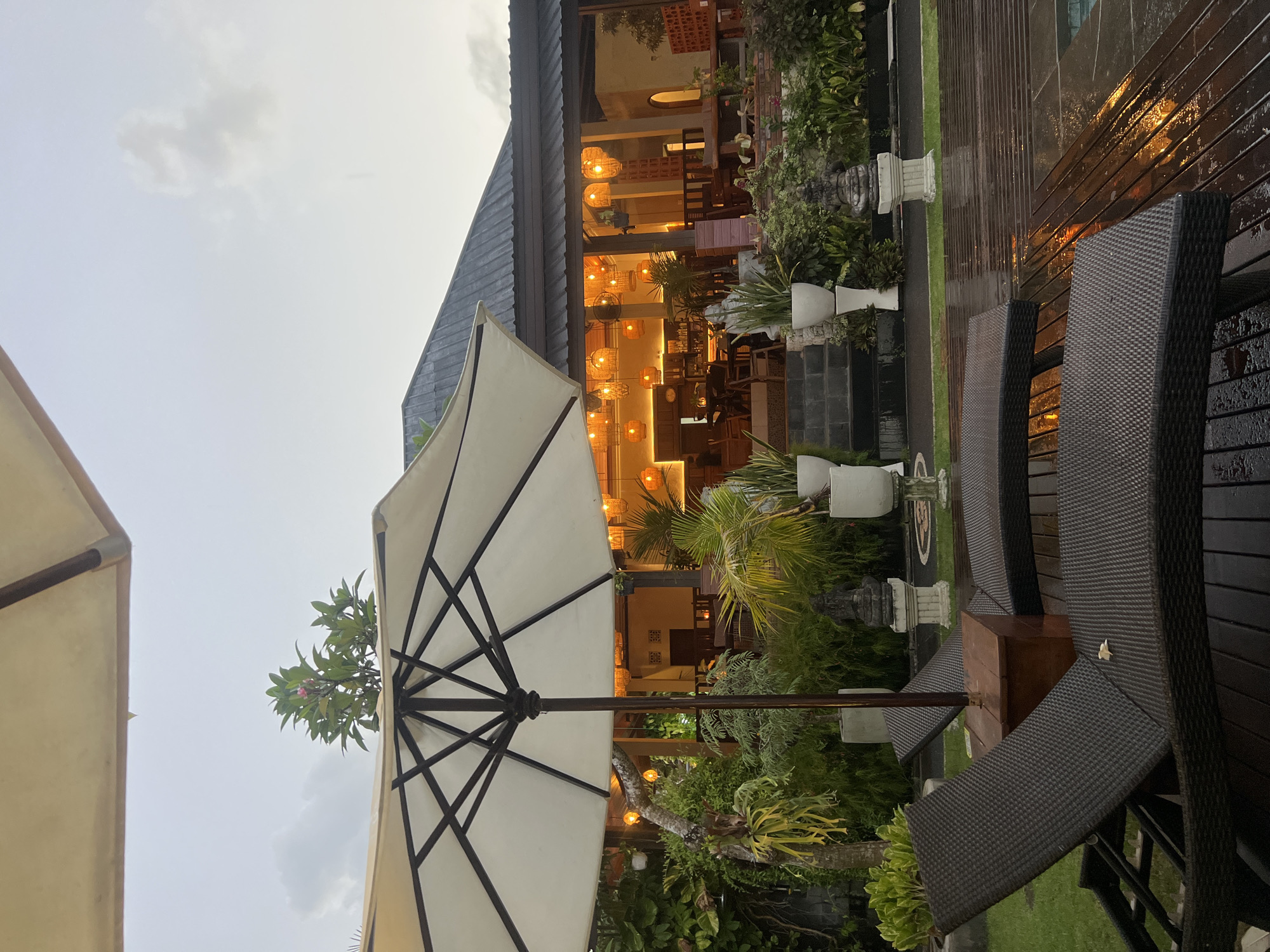 This image depicts an outdoor patio or poolside area, likely part of a restaurant or resort, during dusk or early evening. The scene is dominated by a large, white or cream-colored outdoor umbrella with a black frame, positioned centrally in the foreground.
To the right, several dark brown or black wicker lounge chairs with textured surfaces are arranged on a wet, dark wooden deck. The wetness of the deck and chairs, along with reflections from the artificial lights, suggests recent rain or ongoing light precipitation. Adjacent to the deck is a strip of green lawn and lush tropical foliage, including potted plants and larger trees. A stone statue on a white pedestal is partially visible amidst the greenery.
In the background, a building with a dark-tiled roof is brightly illuminated from within by numerous warm-toned pendant lights and sconces, indicating an inviting interior space, possibly a dining area or bar. The architecture appears contemporary with open sections, allowing views into the well-lit interior.
The sky overhead is overcast and muted, consistent with twilight or an impending evening storm. No people are visible in the image. The overall atmosphere is tranquil and suggests a comfortable setting for relaxation or dining in a tropical environment, consistent with the location context of Kuta Selatan, Indonesia. There is no visible text in the image.