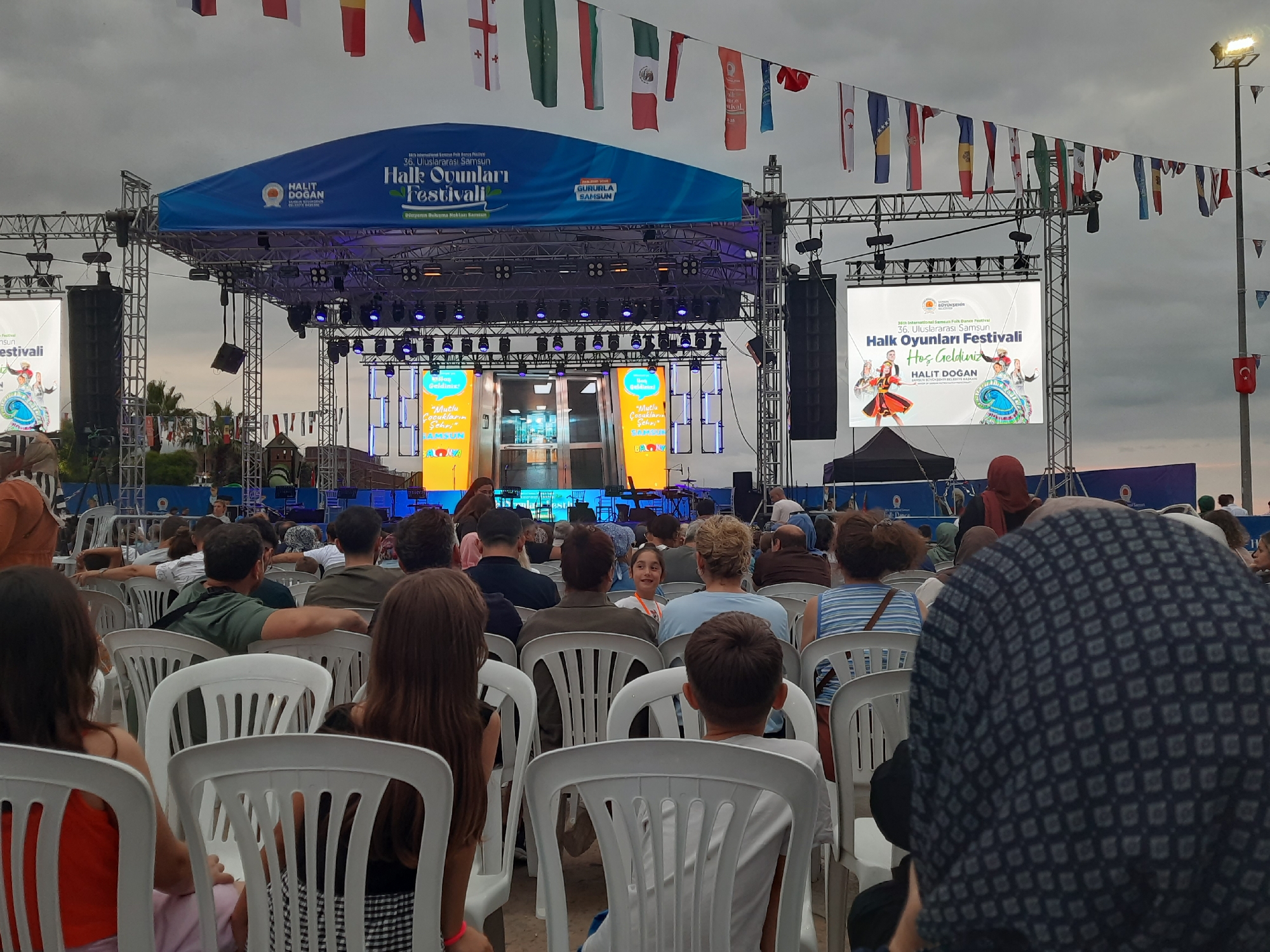 The image depicts an outdoor event at the Halk Oyunları Festivali in Atakum, Turkey. A large stage with a blue banner that reads "HALK OYUNLARI FESTIVALI" is set up, featuring a large screen displaying promotional graphics. The festival appears to be in progress, with numerous people seated in white plastic chairs facing the stage. Flags of various nations are strung across the top of the frame, suggesting an international gathering. In the foreground, the backs of several attendees are visible, some of whom are children. The overall atmosphere appears to be that of a cultural festival or performance.