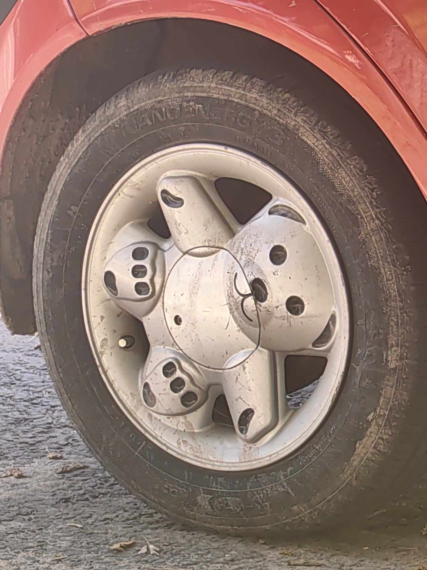 A close-up, eye-level shot shows the front passenger-side wheel and fender of a red car parked on a gravel surface. The wheel has a silver rim with a central hubcap designed to resemble a teddy bear's face, complete with two black circles for eyes and a black, smiling mouth. Five smaller black circles are arranged around the hubcap, possibly representing ears or decorative elements. The tire is dirty, with mud and debris adhering to its sidewall and tread. The car's red fender is also smudged with dirt. The ground is covered in dark gray gravel, with a few dry leaves scattered on the surface. The overall scene suggests the car has been driven on unpaved or muddy roads. This image was taken in the city of Tver, Russia.