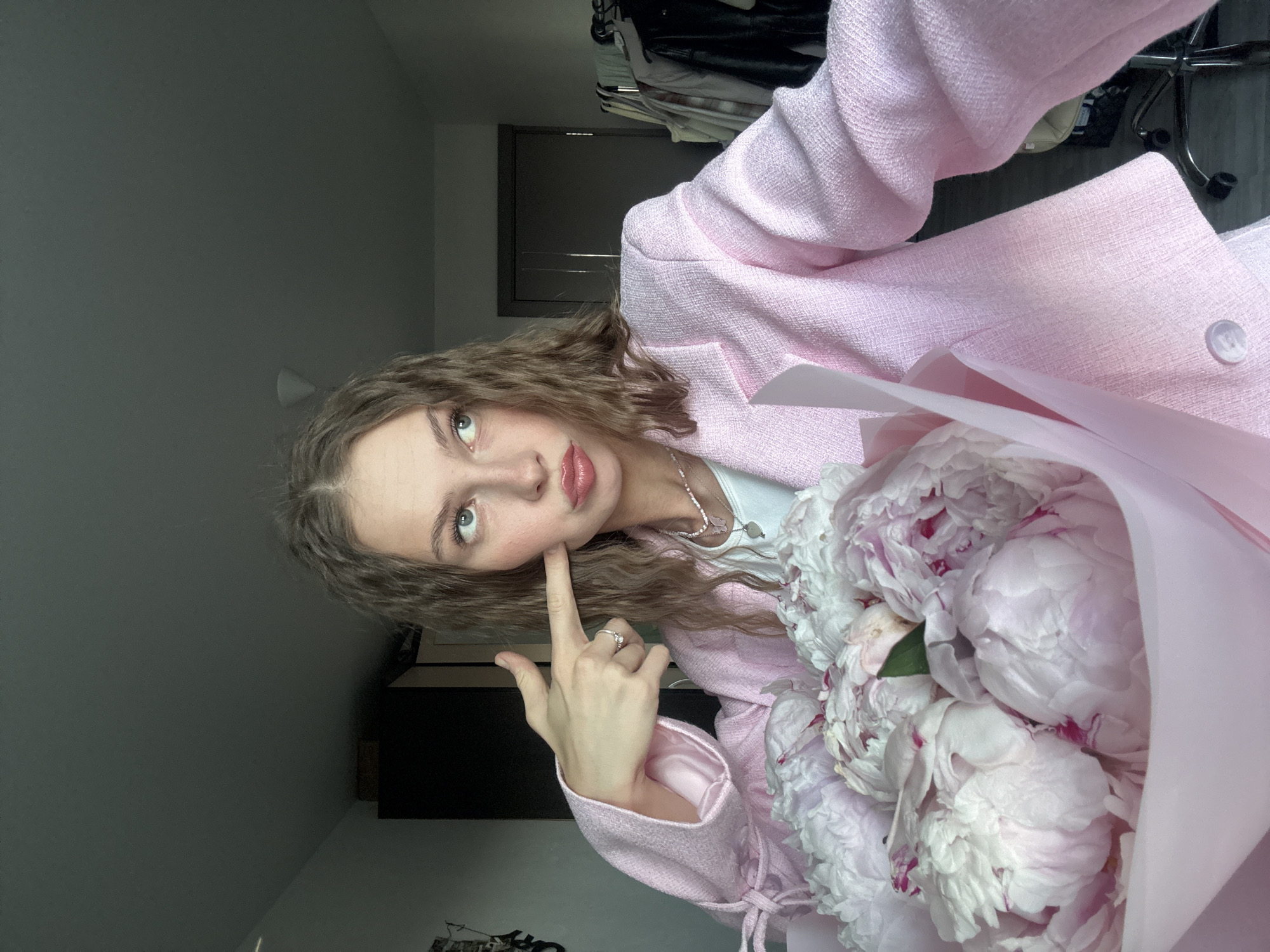 A young Caucasian woman with long, wavy brown hair and blue eyes is holding a bouquet of pink peonies and posing for a selfie in what appears to be a bedroom or dressing area. She is wearing a light pink tweed blazer over a white top and a beaded necklace. Her right index finger, adorned with a ring, is pointed at her cheek. The background includes a dark wardrobe, a doorway, and clothing racks with garments. The scene is lit with natural light from an unseen source, creating shadows on the walls and ceiling. The location is identified as Perm, Russia.