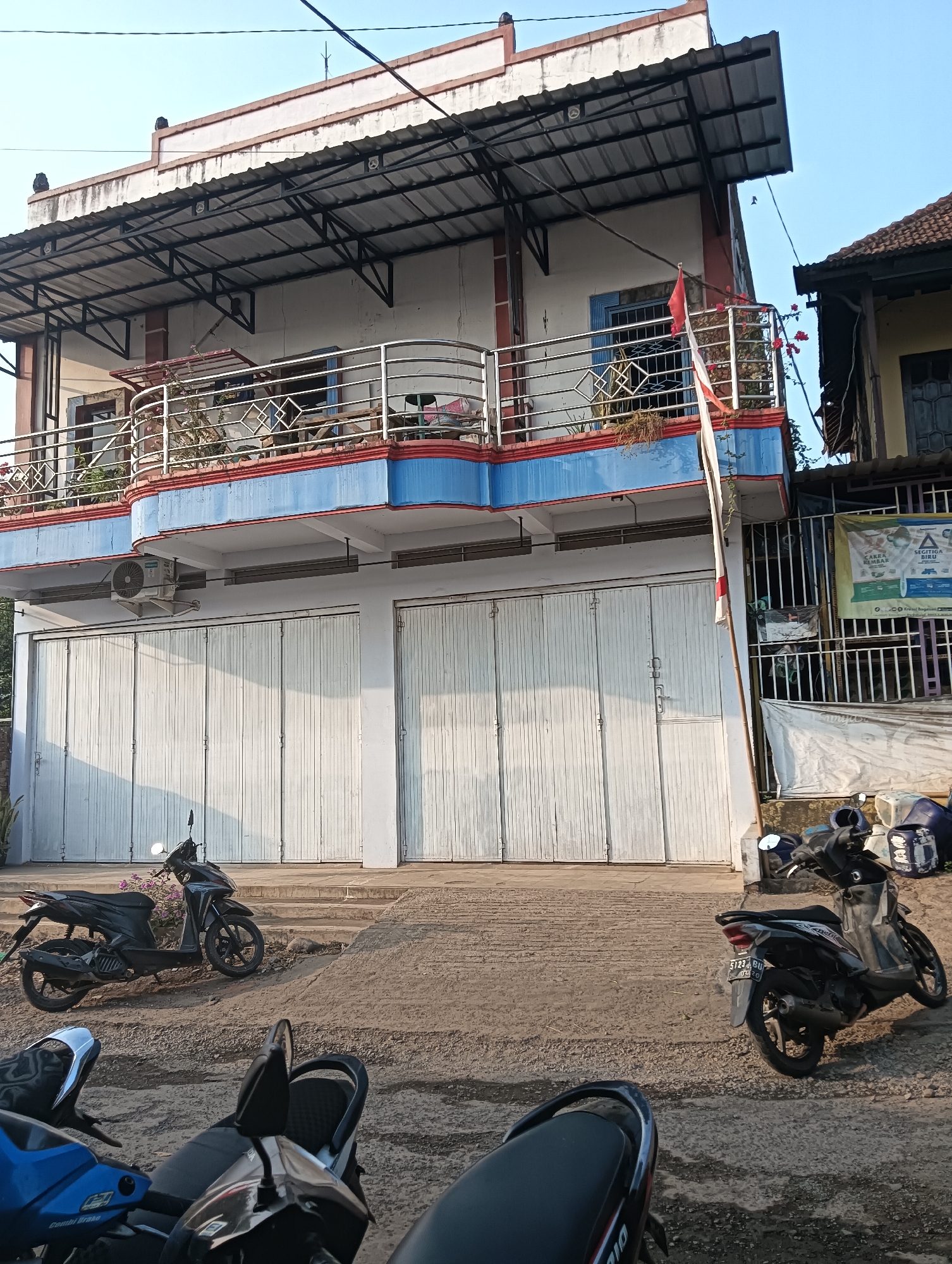 The image depicts a street scene in Cluwak, Indonesia, focusing on the facade of a two-story building and several parked motorcycles. The building has a white and blue color scheme on its upper floor, with a metal awning extending over the ground level. The ground floor is dominated by large, white, corrugated metal roller doors, suggesting commercial use. An air conditioning unit is visible mounted on the building's exterior wall above the roller doors. A flagpole with an Indonesian flag is positioned to the right of the building's entrance.
In the foreground and mid-ground, multiple motorcycles are parked on a paved and gravel surface. Two black scooters are prominent in the mid-ground, one to the left and another to the right. Partially visible in the foreground are parts of other motorcycles, including blue and black models. The overall environment suggests a sunny day, with shadows indicating the time of day. A narrow street or sidewalk is visible in front of the building, and a fence runs along the right side of the property, behind which a banner with text and graphics is partially visible. The architecture appears to be typical of commercial buildings in urban or semi-urban areas of Indonesia.