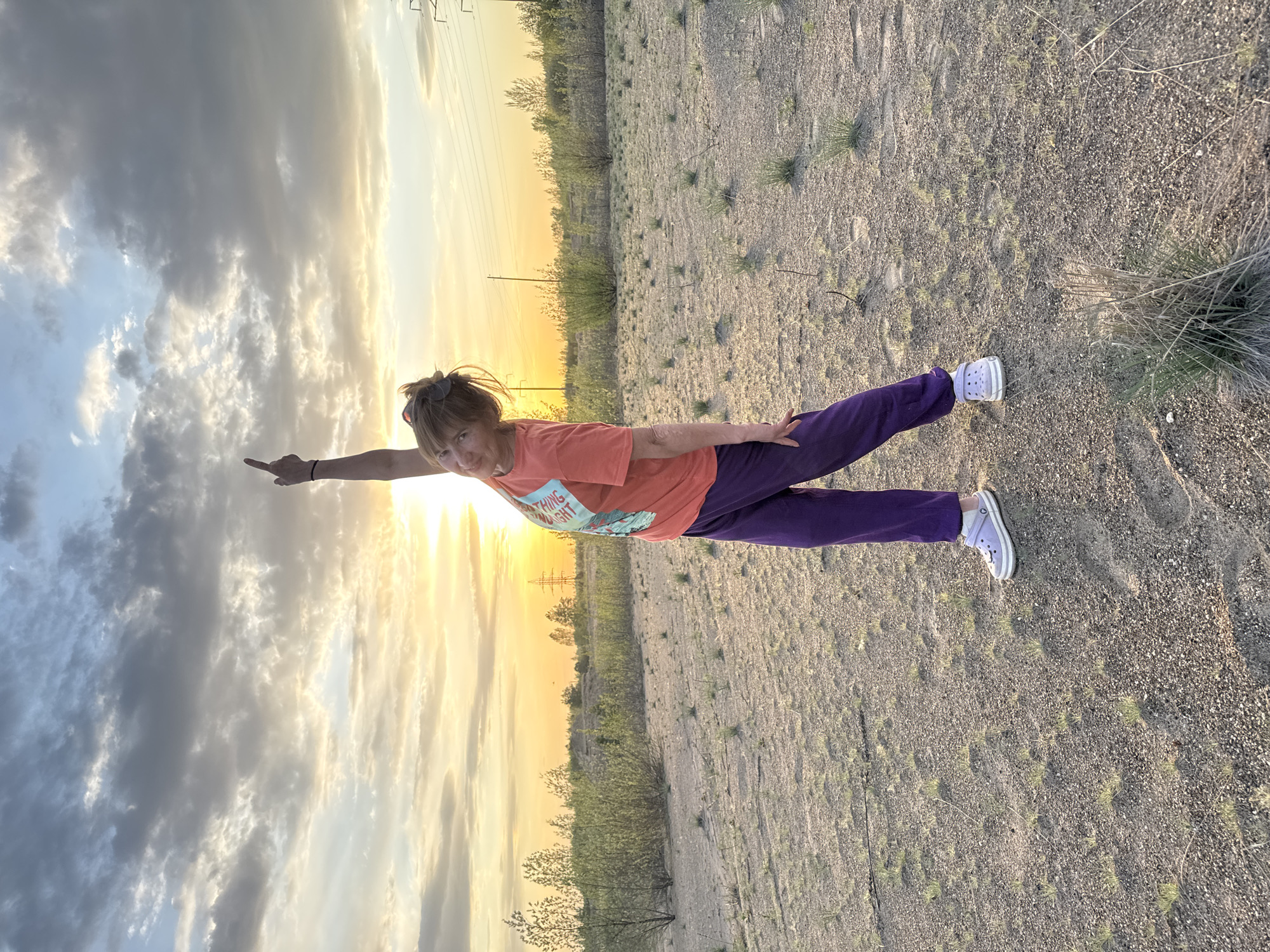 A woman wearing a peach t-shirt and purple pants is outdoors in a field. She is lying on her stomach with her arms extended, appearing to fly like Superman. She has brown hair and is wearing light purple shoes. The field has sparse vegetation. The background includes a yellow sunset and cloudy sky.