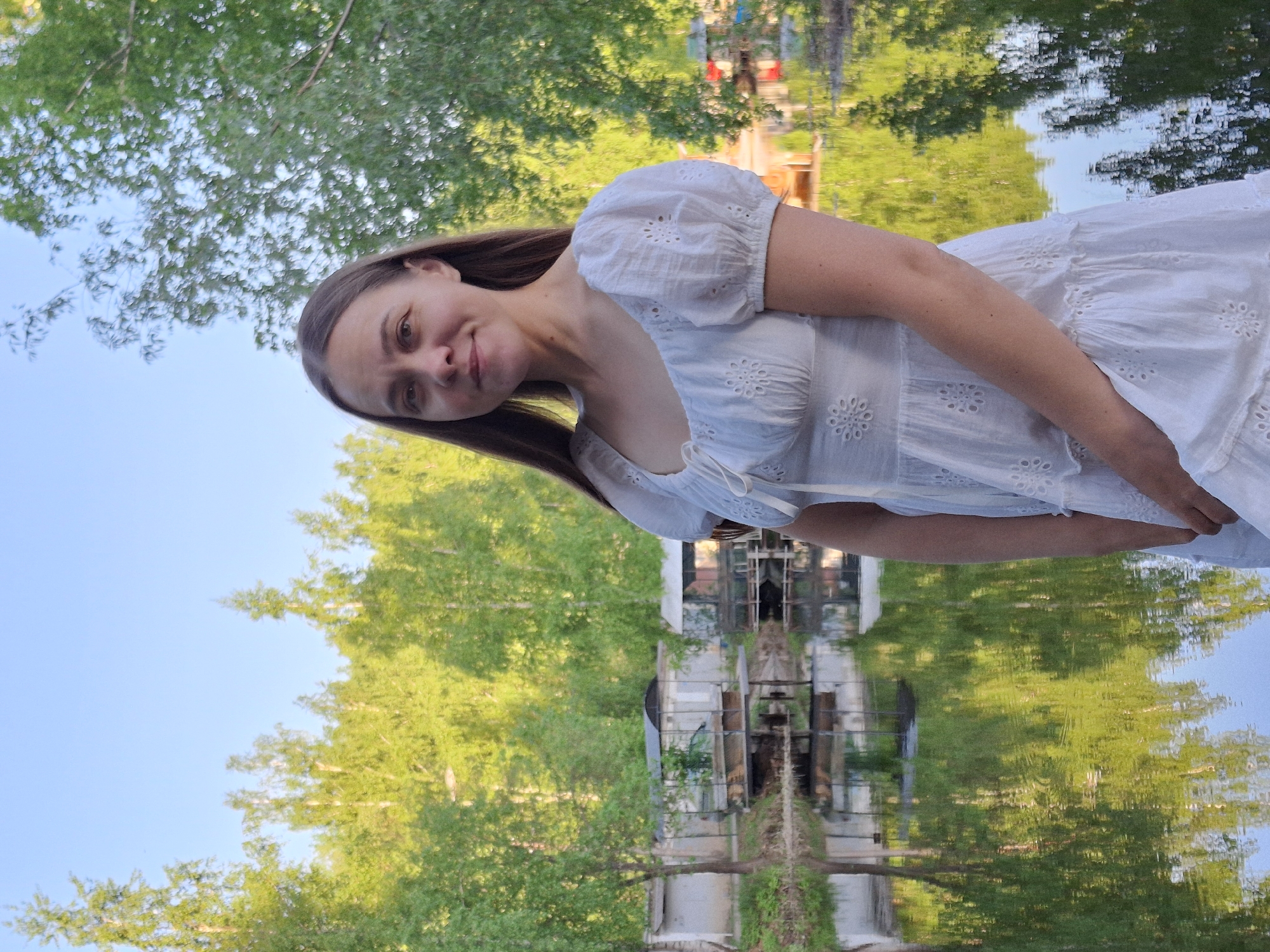 A young woman in a white eyelet dress stands in a park. The location is Chaikovsky, Russia. The background shows trees and a building. The image seems to be reflected in water, suggesting it was taken near a pond or lake.