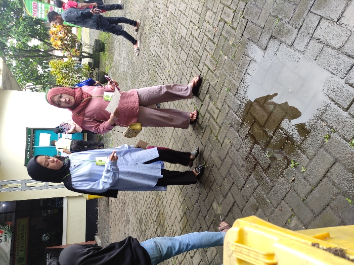 The image depicts an outdoor scene in Kepanjen, Indonesia, featuring several individuals gathered on a paved area made of interlocking bricks. The ground appears wet, with a prominent puddle reflecting the sky and surrounding elements, suggesting recent rain.

In the mid-ground, two women wearing hijabs and long-sleeved tops are central. The woman on the left wears a light blue top and a black hijab, along with black pants and sandals. She has a green lanyard with an ID card around her neck. The woman on the right wears a pink top, a matching pink hijab, and light brown pants, with sandals and a yellow shoulder bag. She is holding a piece of paper and also has a green lanyard with an ID card around her neck, looking engaged or contemplative.

In the background, a man in a dark shirt and pants is standing, partially obscured by foliage. Another person is partially visible behind him. In the foreground, the legs of a person in blue jeans and a dark top are visible next to a yellow object, possibly a barrier or container.

The setting appears to be outside a building, with greenery visible in the background. A sign on a green banner in the upper left of the image (when oriented correctly) is partially visible. Legible text includes "DINAS KESEHATAN" (Department of Health), "KEPANJEN", "PUSKESMAS" (Public Health Center), and "GRATIS" (Free). This indicates the location is likely a public health center and suggests an event or service is being offered there. The time of day is daylight, under possibly overcast conditions given the flat lighting and wet ground. The overall mood is casual, with people standing and possibly waiting or interacting.