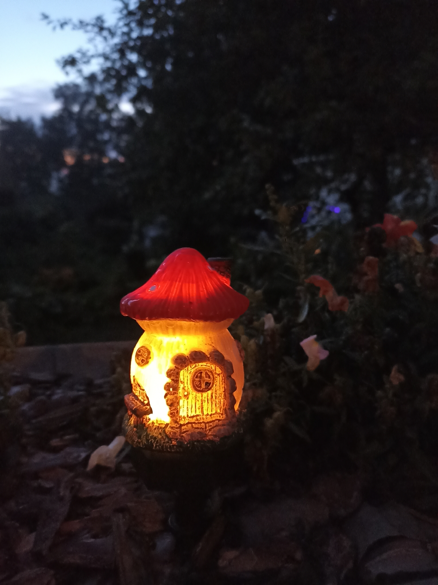 In the city of Mochische, Russia, an illuminated mushroom-shaped garden ornament sits on a dark, bark-covered ground. The ornament, made of ceramic, has a bright red cap and a yellow-lit interior, featuring a detailed door with a circular window. Surrounding the ornament is dense foliage, with some blurred flowers visible in the background. The scene is set at dusk, with a twilight sky visible above the trees.