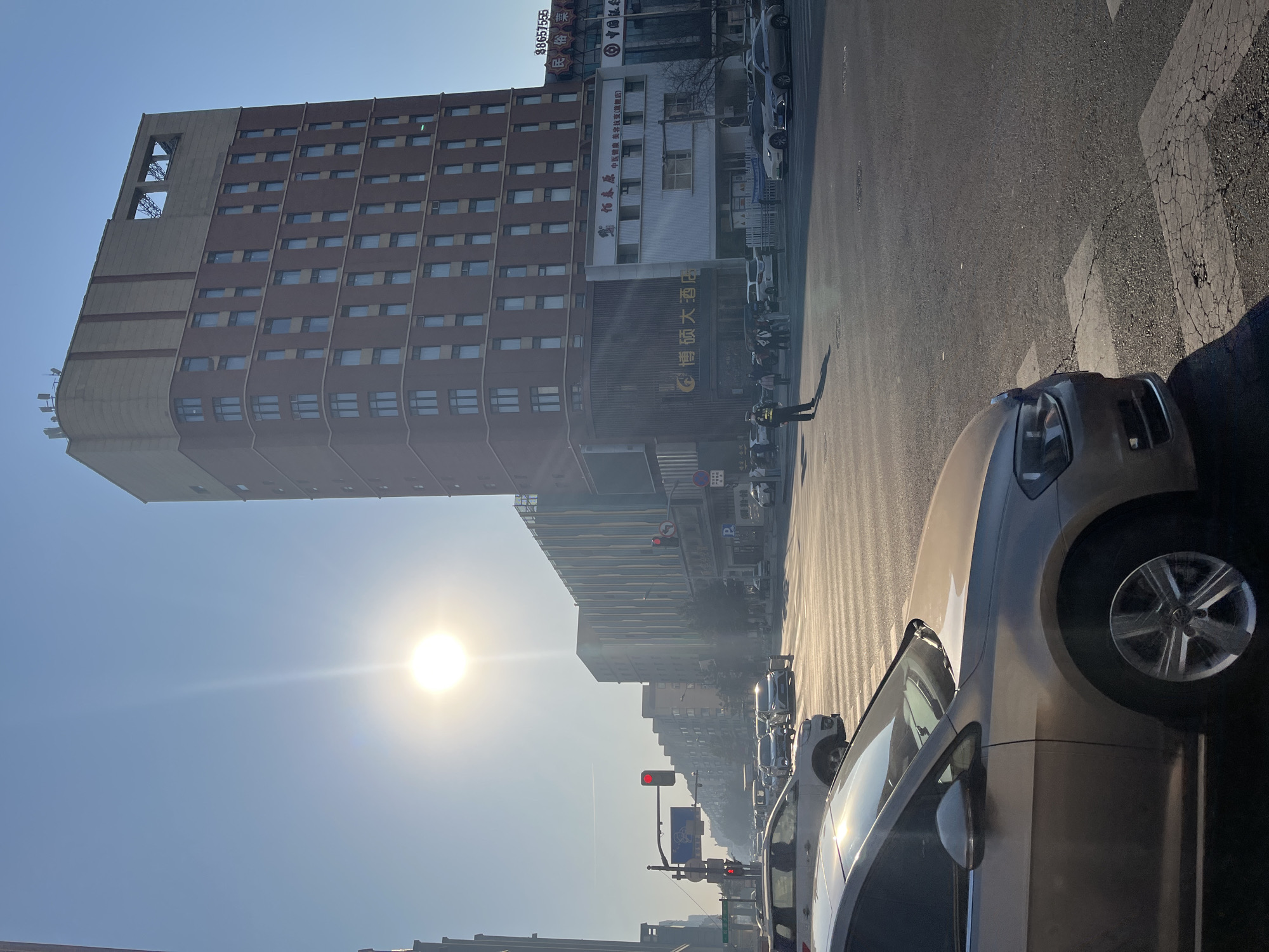 The image shows a street scene in Changchun City, China. The sky is bright and blue. A tall, boxy building with many windows stands on the left side of the frame. Adjacent to it is a shorter, lighter-colored building with signs in Chinese characters, including "博硕大酒店." Several cars are parked along the right side of the street and in the distance, suggesting traffic. A person in uniform stands in the street near the hotel. The sun is shining brightly, creating glare and shadows.