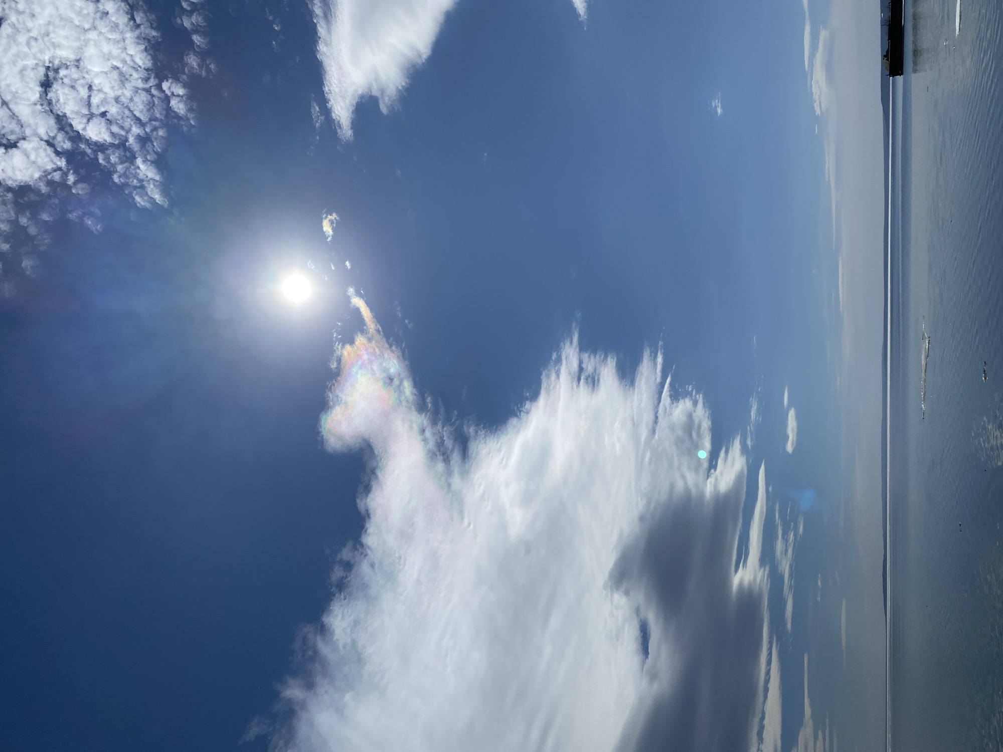 The image depicts a daytime scene with a bright sun in a blue sky. Various types of clouds are present, including cumulus and cirrus formations. A body of water is visible on the right side of the frame, suggesting a lake or river. The horizon line is distinct but somewhat hazy. The location in the city of Ulyanovsk, Russia, is not explicitly discernible from the visual information provided. No people or specific objects indicating urban landmarks are visible. The environment is primarily natural, consisting of sky, clouds, and water.