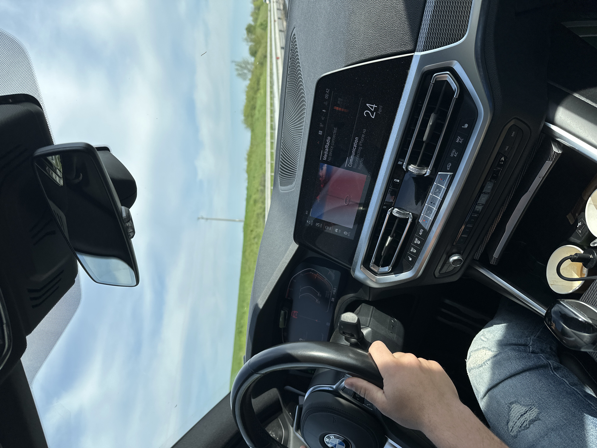 The image captures a first-person perspective from the driver's seat of a modern car, likely a BMW given the steering wheel logo. A person's right hand is visible on the steering wheel, and a leg in ripped blue jeans is also seen.
The interior of the car is prominent, featuring a digital dashboard and infotainment system. The infotainment screen displays "09:42" for the time, "24 April" for the date, and messages indicating "Media/Radio No device connected." and "Communication Reconnecting iPhone на Raycho." The instrument cluster shows "D8" and an outside temperature of "+16.5" degrees Celsius. Below the infotainment screen, physical climate control buttons are visible, including "AUTO", "MENU A/C", and "MAX A/C". To the right, a charging cable is plugged in, and a paper cup sits in a cup holder.
Through the windshield, a clear blue sky with scattered white clouds is visible, suggesting good weather conditions. The landscape outside appears green and open, possibly a highway or rural road with trees and grass lining the route. The rearview mirror is also visible, reflecting the sky.
The activity depicted is driving, with the driver's hand on the wheel. The visible text includes the time, date, car system messages, and control labels, as noted above. The provided context indicates the location is Voyvodovo, Bulgaria.