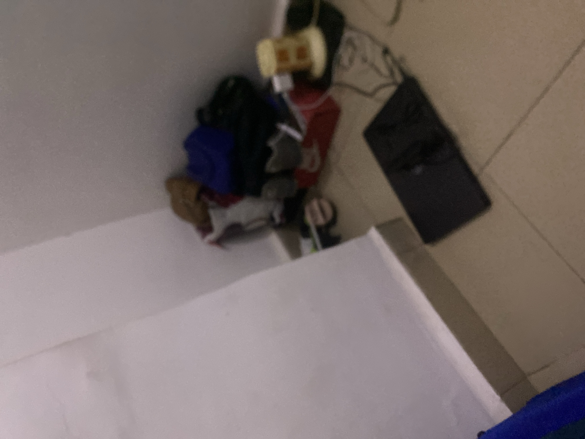 The image depicts a corner of a room, possibly in Howayi, Nigeria, characterized by a cluttered floor and two walls meeting at a right angle. The left wall is a plain, light-colored surface, while the floor is tiled in a neutral tone. In the corner, a pile of various items is visible, including what appears to be clothing or bags in shades of blue, black, and brown, as well as a red bag or box. A yellow object with a segmented pattern, possibly a container or light fixture, sits amongst the clutter. To the right of this pile, a dark, flat electronic device, likely a tablet or laptop, lies on the floor, with charging cables connected to it. The scene is dimly lit, and the focus is soft, suggesting a casual or storage area rather than a formal setting.
