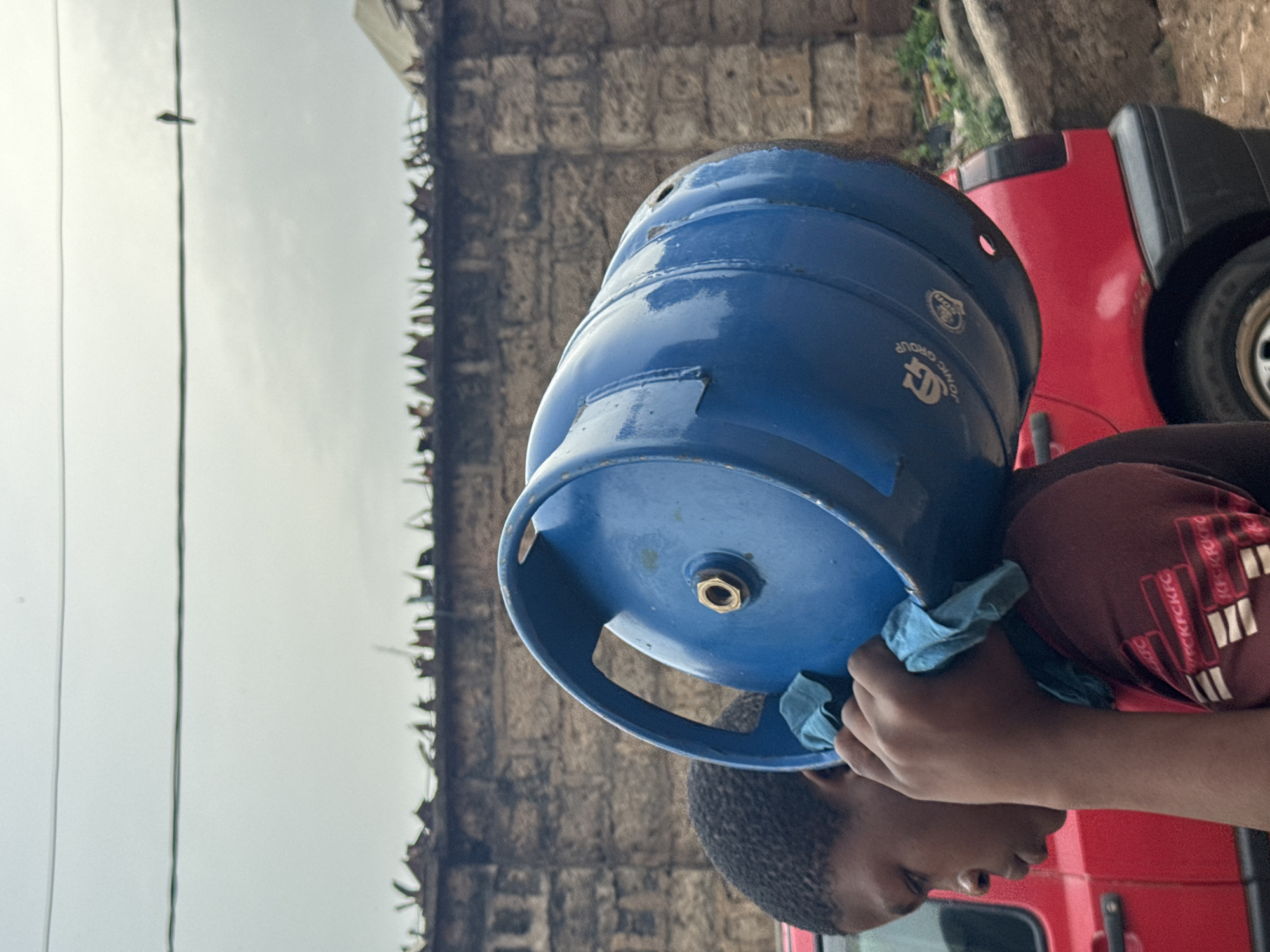 A person carries a blue gas cylinder on their head in Onitsha, Nigeria. The cylinder is slightly damaged. The person wears a maroon shirt. A red vehicle is visible behind them. A wall with a metal security fence sits in the background. Overhead wires are visible against a gray sky.