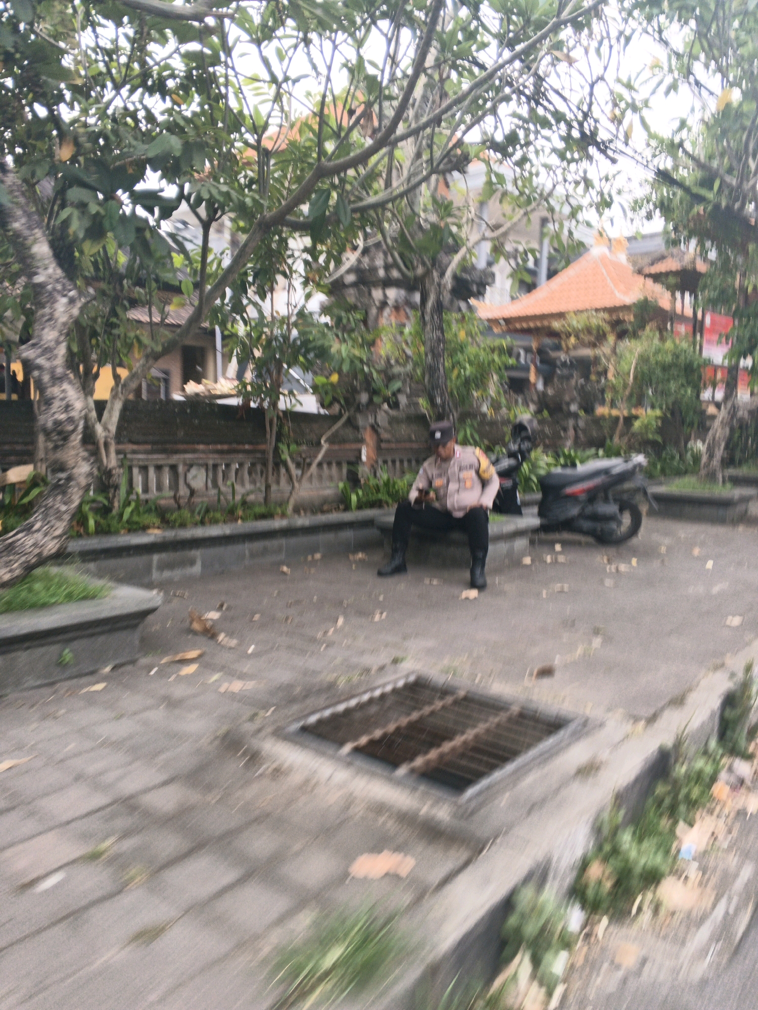 In Ubud District, Indonesia, a police officer in uniform is seated on a low stone wall, looking at a smartphone. The officer is wearing a light-colored shirt with patches and dark pants. A black scooter is parked behind the officer. The scene is an outdoor paved area with trees, foliage, and a building with an orange tiled roof in the background. There is a metal grate set into the pavement. The image has motion blur, suggesting it was taken from a moving vehicle.