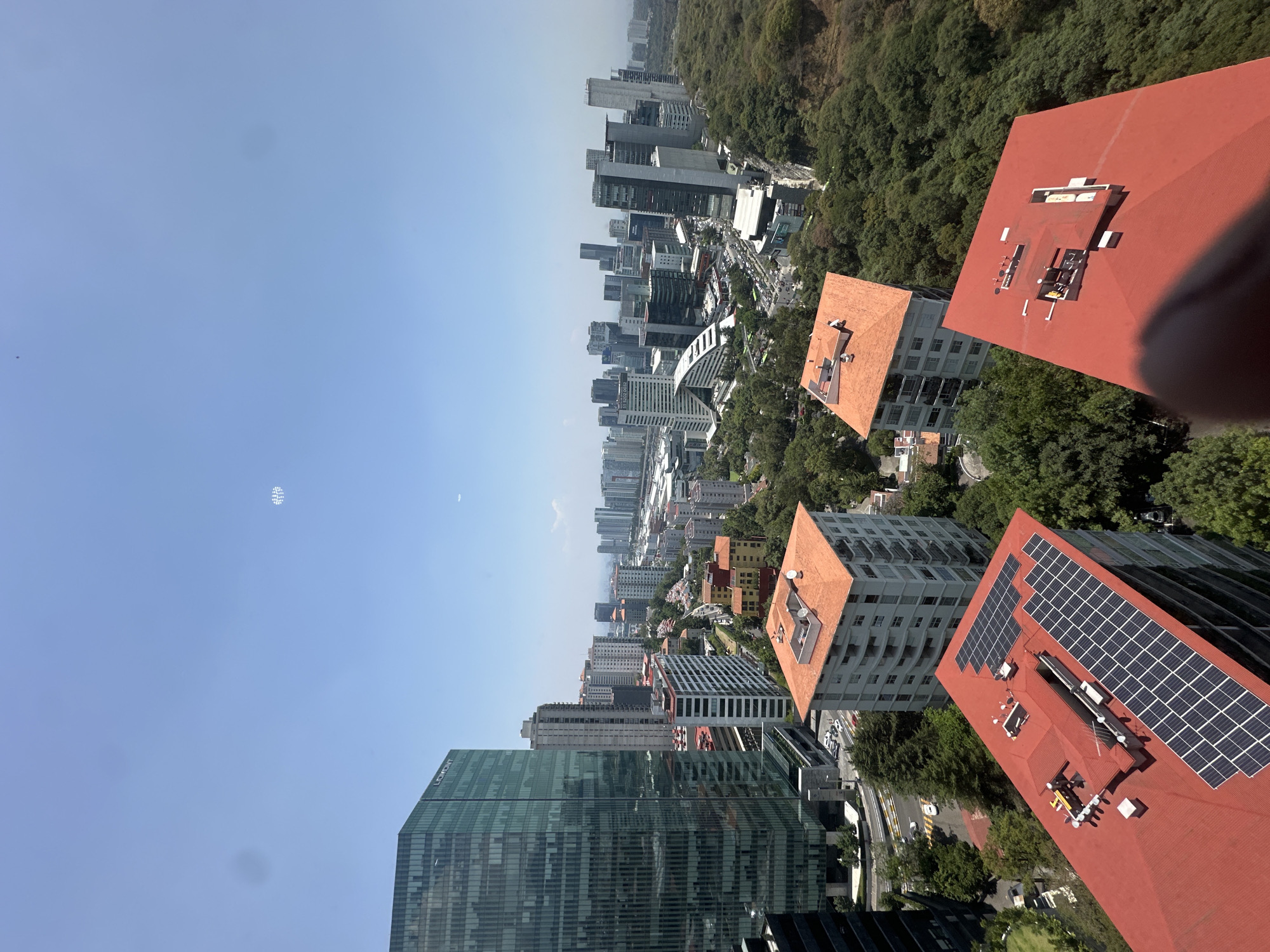 This is a high-angle, outdoor shot of a cityscape in Mexico City, Mexico, taken during the daytime. The view is looking down from a high building, capturing several buildings, some with red tile roofs and some with solar panels, and other modern high-rise structures against a backdrop of trees. The sky is blue, with a contrail visible in the upper left.
