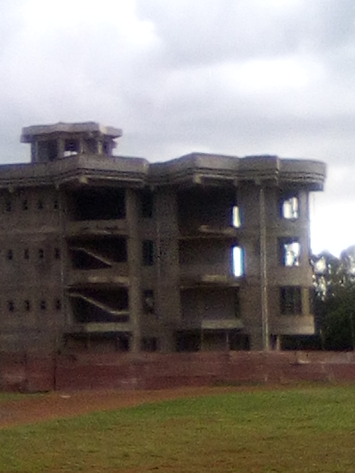 The image displays an unfinished, multi-story concrete building located in Bondo, Kenya, under an overcast sky. The structure features a complex, angular design with multiple sections and levels, including a distinctive rooftop component. Numerous window and door openings are visible throughout the building, most appearing unglazed. On the left side, two interior staircases are discernible through open sections. A low, reddish-brown wall separates the building from a foreground area composed of green grass and reddish-brown earth. No individuals, vehicles, or active construction equipment are present in the scene. Dense green foliage is visible to the right of the building, partially obscured by the structure.