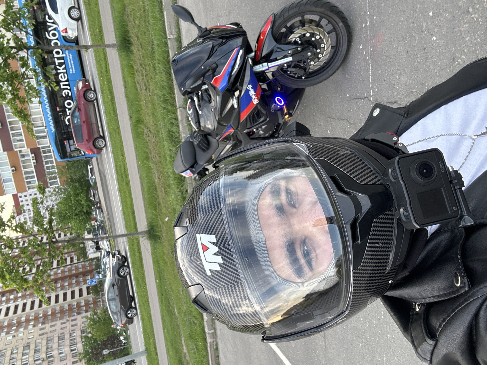 A high-angle selfie shows a person wearing a black carbon-fiber motorcycle helmet with a clear visor. Mounted on the side of the helmet is a black action camera. The person is wearing a black leather jacket over a white garment and a silver necklace is visible.
Behind the person is a motorcycle with red, blue, and black colors. A blue LED light is visible near the front wheel. The background shows a cityscape. In the background, cars and a bus with the partially visible text "это электробус" (this is an electric bus) are on the street. Based on the bus text and general appearance, the location is likely Moscow, Russia. Buildings are visible in the background. Grass and trees are also present. The setting appears to be outdoors during the daytime.