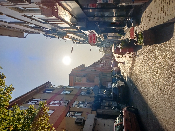 The image shows a narrow street in Istanbul, Türkiye, located at 40.962 latitude and 28.825 longitude. The street is lined with buildings on both sides, with cars parked along the edge of the street. The street is paved with cobblestones and there are a few potted plants along the side. The sun is shining brightly in the sky, casting a warm glow over the scene. A single person walks down the middle of the street, heading towards the sun. To the left, a red car is parked near the beginning of the street, facing the opposite direction of traffic. In the center of the image are several parked cars, some in front of shops and others at the curb. To the right, the image features several buildings that are all multi-story. There is a sign in the window of one building that reads "ÇATI CAFE." The sign is in Turkish and refers to the roof of the building.