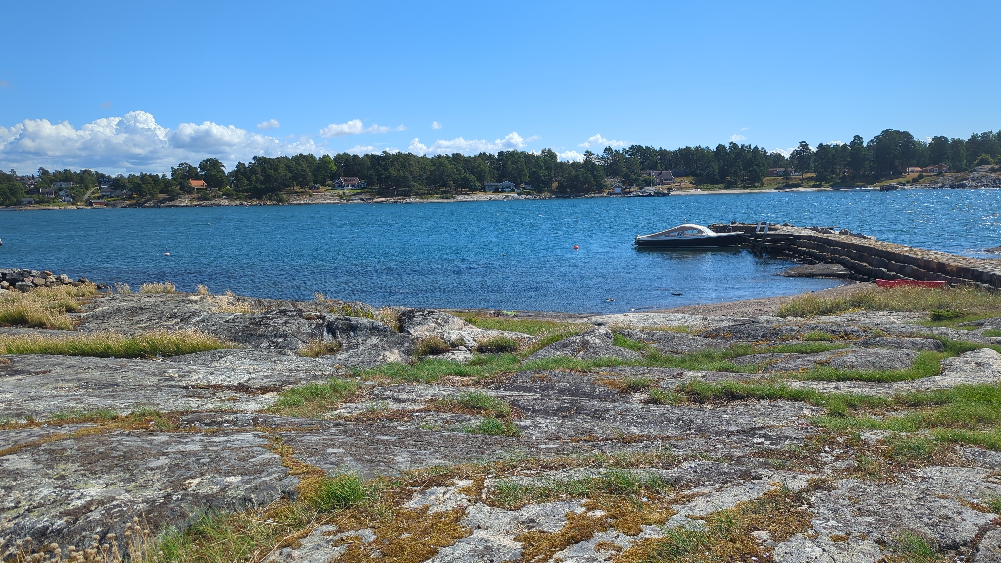 A tranquil, sun-drenched day unfolds at the coastline of Larkollen, Norway, depicted in this landscape photograph. The foreground is dominated by weathered, grey rock formations dotted with patches of dry grass and moss, suggesting a rugged, natural terrain. Beyond this rocky expanse, a calm, azure sea stretches out, its surface gently rippled by a light breeze.
On the left side of the water, a densely wooded shoreline is visible, punctuated by several scattered residential buildings with characteristic red and orange roofs. These houses appear nestled amongst the trees, hinting at a peaceful, suburban or rural setting. A thin line of beach separates the water from the treeline.
To the right, a substantial stone pier juts out into the sea. Moored to this pier is a sleek, modern white and dark-hulled boat, its polished surface gleaming in the sunlight. Beside the pier, a bright red object, possibly a kayak or a small dinghy, rests on the shore. Further out in the water, a small, orange buoy floats, and a white buoy is visible closer to the left shore.
The sky above is a brilliant, clear blue, with a few fluffy white clouds drifting lazily across the upper portion of the frame, adding to the idyllic summer atmosphere. The overall impression is one of serene coastal beauty and a quiet, relaxed day.