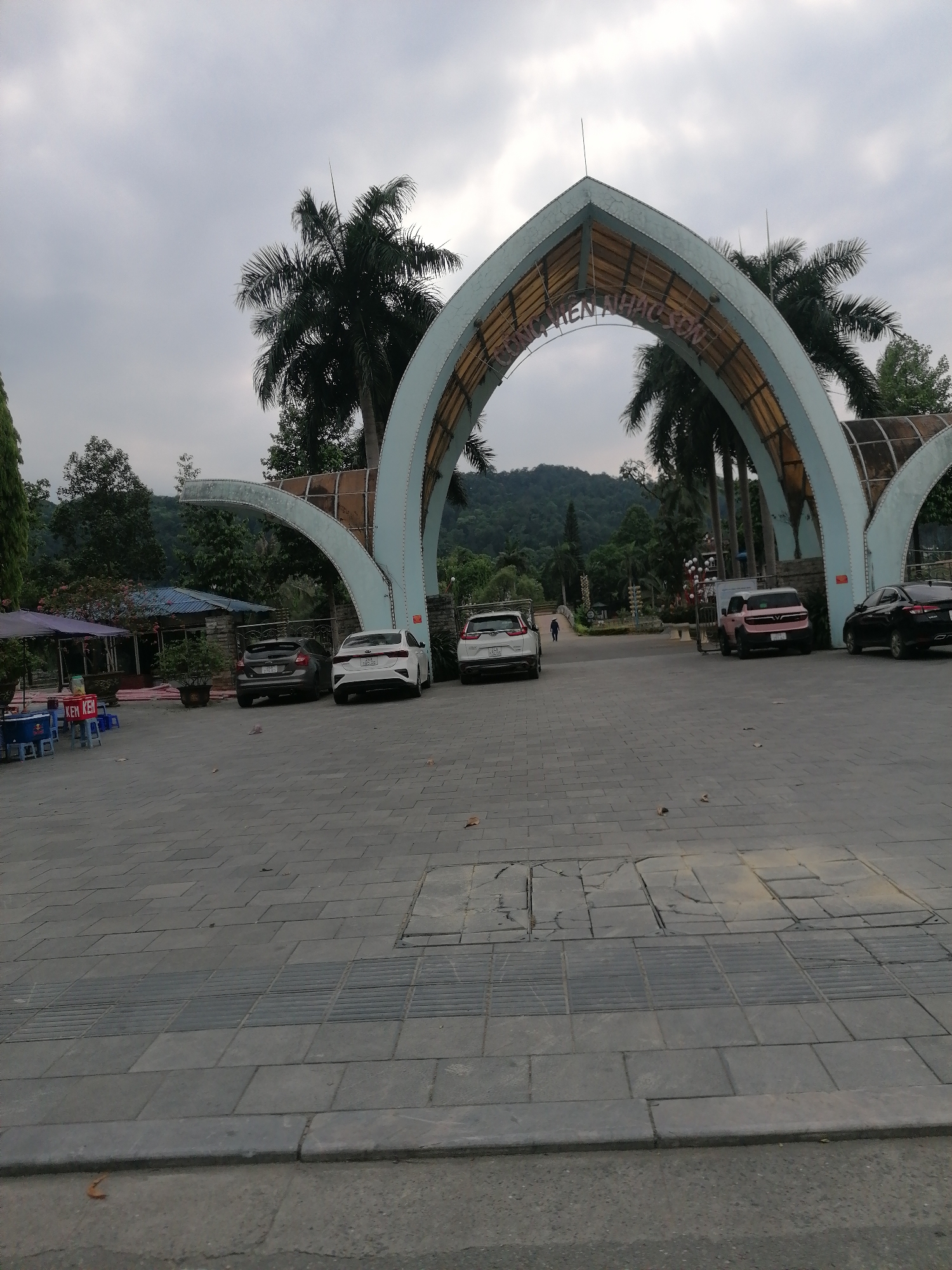 The image shows the entrance to a park or recreation area in Lao Cai Ward, Vietnam. A large, ornate archway spans the entrance road, with the inscription "Cong Vien Nhac Son" visible. Several cars are parked along the roadside near the entrance. Behind the entrance, a lush landscape of trees and hills can be seen. The foreground is dominated by a paved area with discernible tactile paving strips. To the left of the frame, there is a small food stall with tables and chairs. A person is walking on the road further inside the park. The sky is overcast.