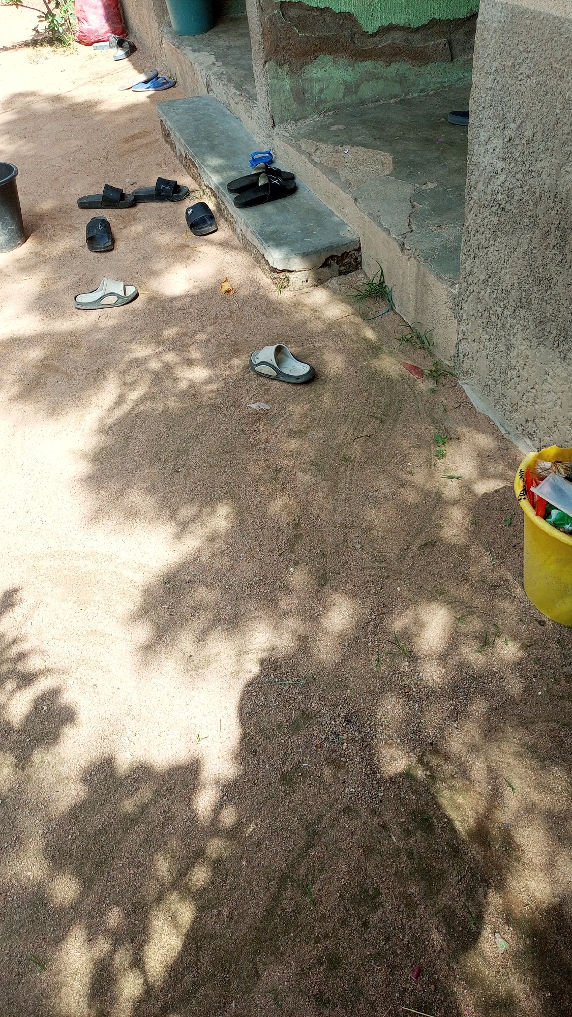 The image depicts an outdoor scene with several pairs of sandals and flip-flops scattered on sandy ground in front of a building. The building has a concrete step and wall with some green and brown paint. In the background, a red bag and a blue bucket are visible. To the right, a yellow bucket filled with various items, including what appears to be trash, is placed on the ground. Tree shadows are cast across the sandy area. The scene suggests a casual, everyday environment, potentially a residential or communal outdoor space in Jalingo, Nigeria.