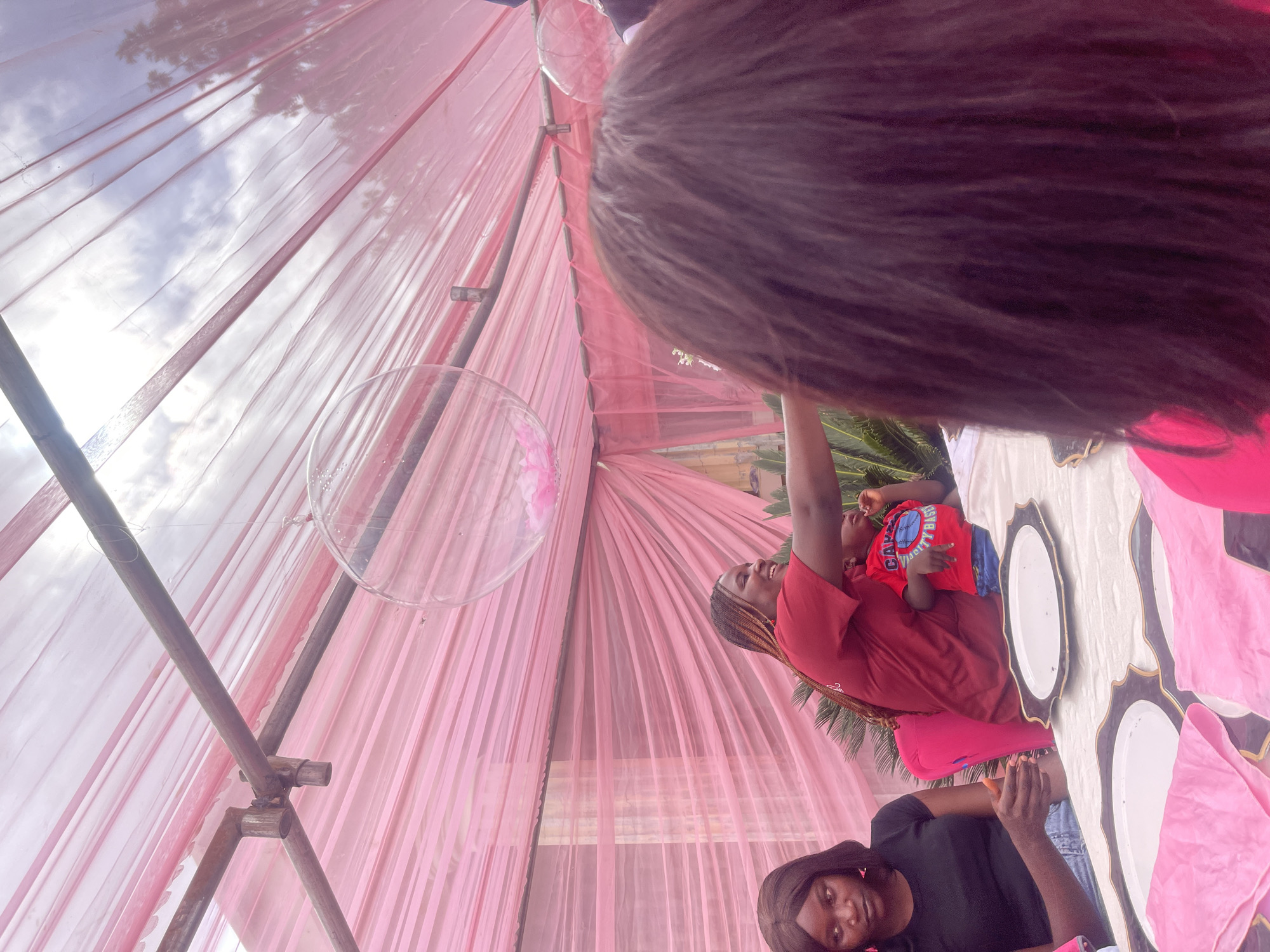 This image depicts an outdoor event under a pink draped canopy supported by metal poles. In the foreground, the back of a person with dark, textured hair is visible. To the left, a woman with dark hair and pink earrings is seated, wearing a black top and blue jeans. She is holding an orange object. Beside her, a woman with braided hair, wearing a red top, is leaning forward and smiling, reaching towards a young child in a red shirt with blue lettering. The child is looking upwards. The scene appears to be a celebration or gathering, possibly a birthday party, given the festive decorations and the presence of young children. The location is specified as Jos, Nigeria, suggesting an urban or semi-urban setting with vegetation visible in the background. The table in the foreground is set with ornate plates and pink napkins.
