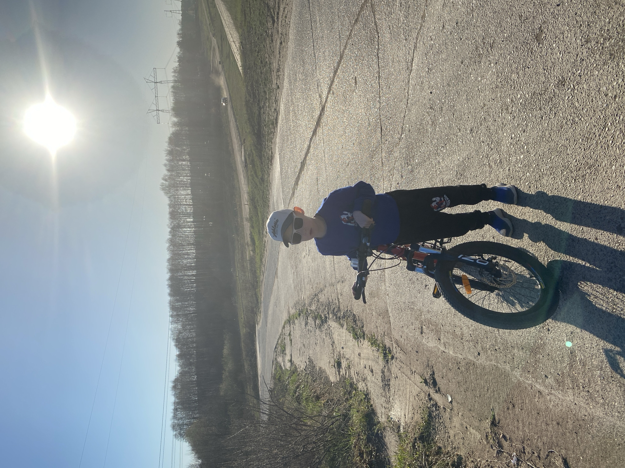 A boy is riding a bicycle on a paved road. He is wearing a blue shirt, black pants, blue shoes, sunglasses, and a white helmet. The road is cracked and there is grass growing along the edges. There are power lines and trees in the background. The sun is shining brightly. The location may be Chaikovsky, Russia, but this cannot be confirmed without additional context.