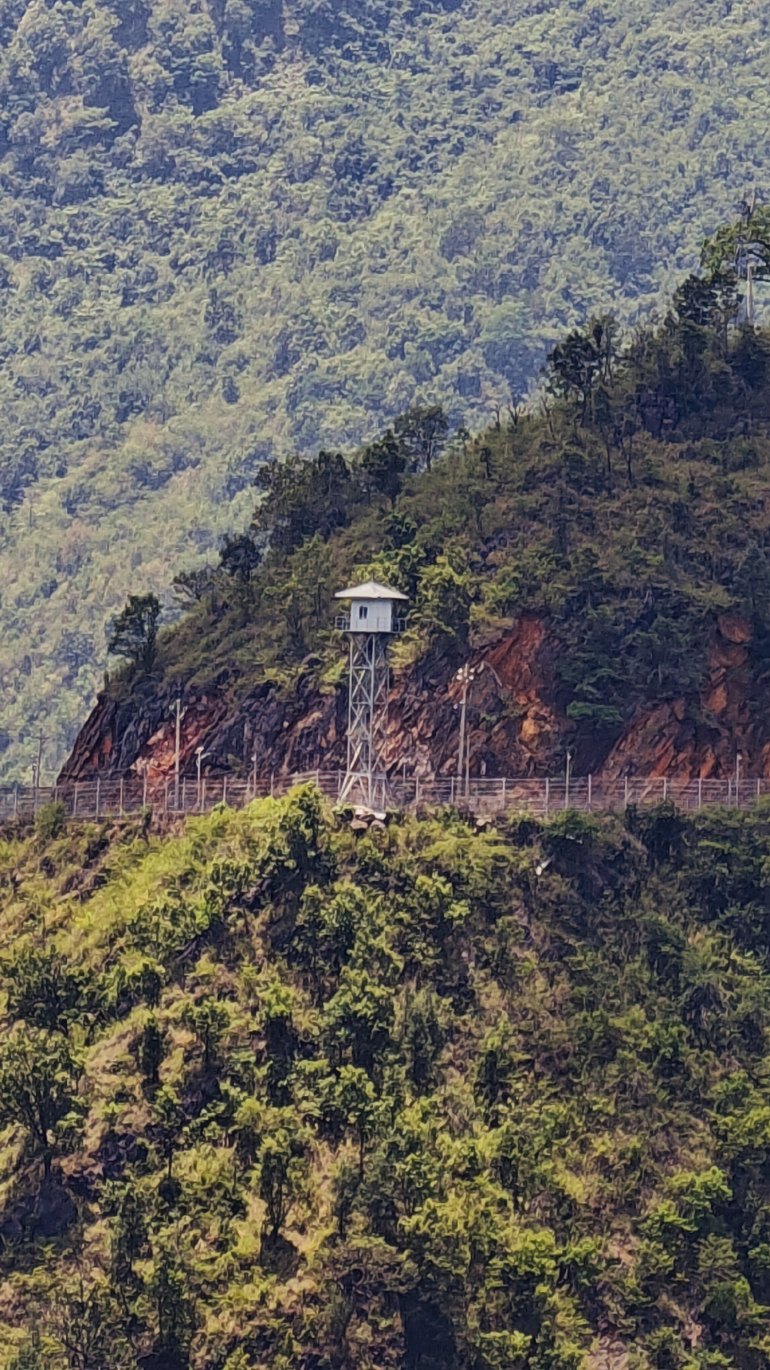 The image shows a lookout tower in Lũng Cú Commune, Vietnam. The tower is a small, white structure with a metal frame, situated on a hillside. A fence runs along the hillside near the tower. The surrounding environment consists of lush, green vegetation and steep terrain. The scene appears to be outdoors and is captured during daylight hours. The image appears to be taken from a distance.