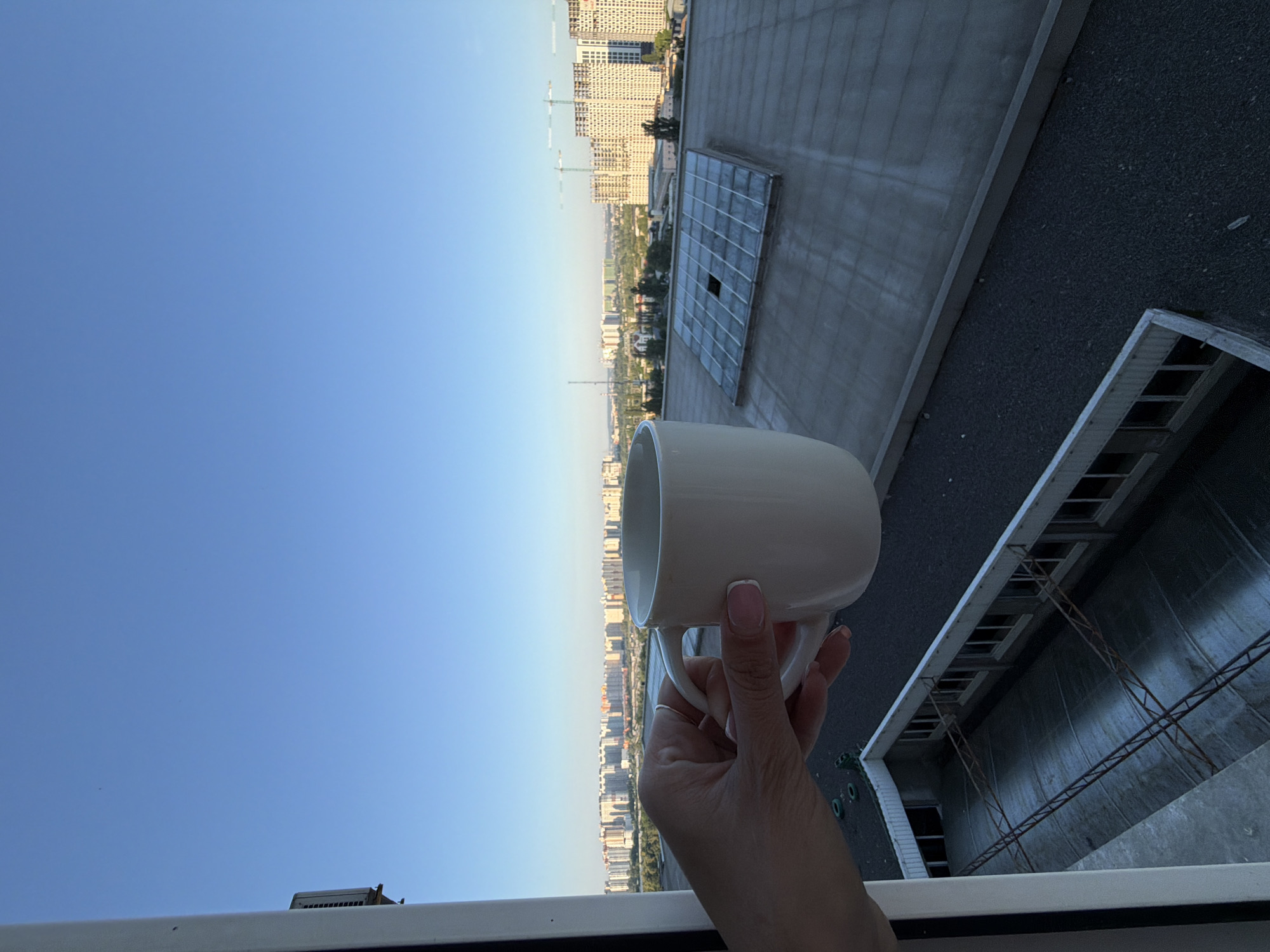 An over-the-shoulder shot shows a person's hand holding a white mug of coffee. The background reveals a cityscape, indicating the scene is likely in Kyiv, Ukraine. In the distance, residential buildings and construction sites are visible under a clear blue sky. In the foreground, the rooftop of a building with a large, tiled skylight is present, along with an adjacent building with a metal-covered roof and industrial-style windows. The image captures a moment of enjoying a morning view from a high vantage point.