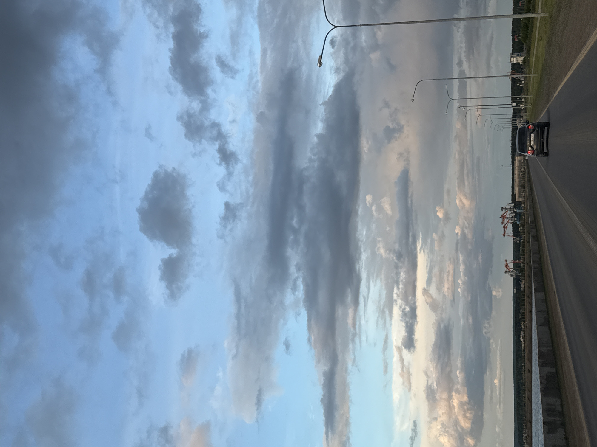 The image shows a road with a car driving away, lined with streetlights. The background is a cloudy sky, with a mix of light blue and dark grey clouds. In the distance, there is a cityscape. Cranes are visible on the right side, as well as an area near water. The image appears to be taken in Chaikovsky, Russia, based on the visual cues of city infrastructure, water feature and cloud formations.