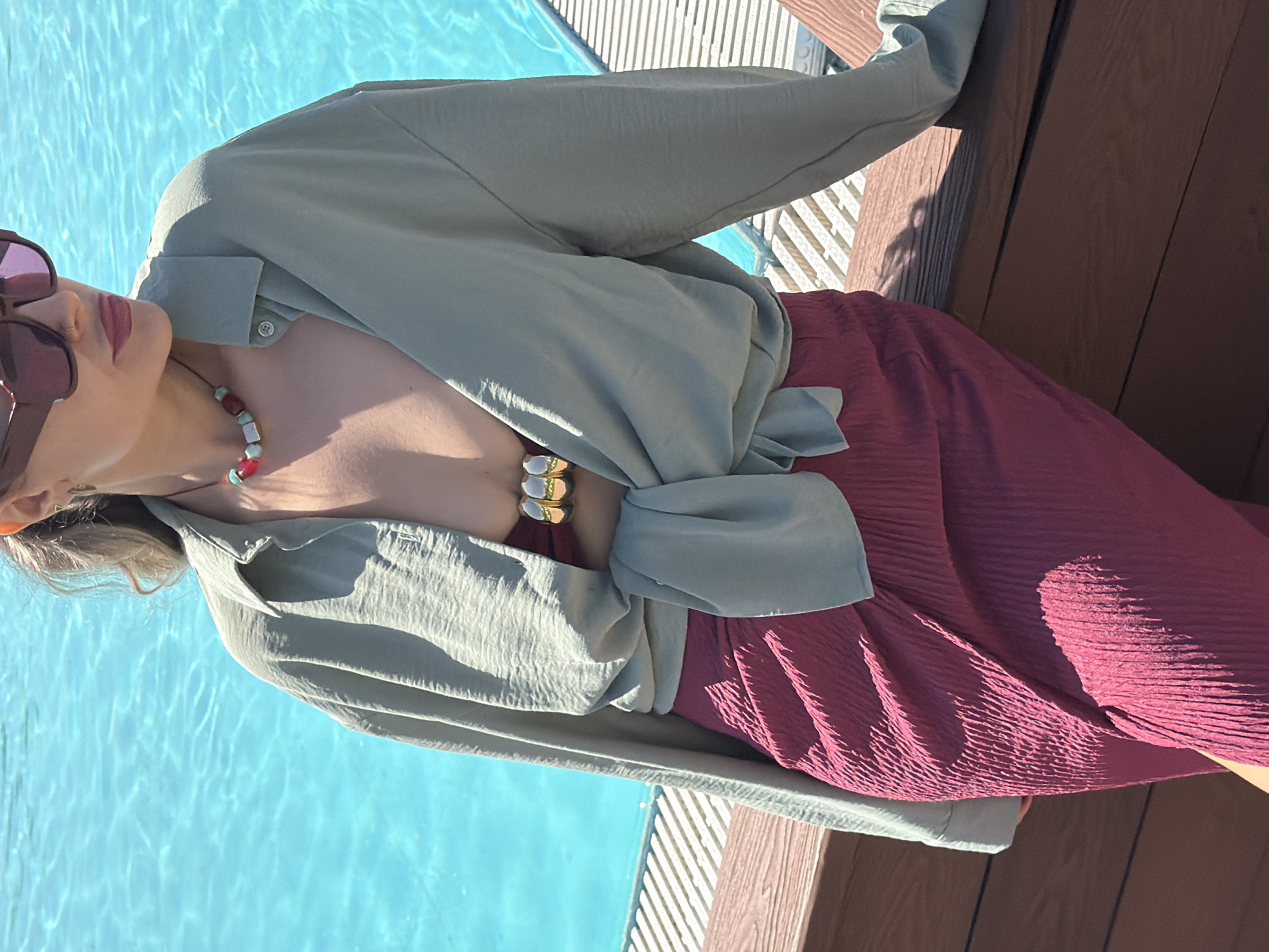 An overhead shot, rotated 90 degrees to the left, captures a person relaxing outdoors by a swimming pool in Krasnoyarsk, Russia. The individual, likely female, is wearing large, dark reddish-purple sunglasses and a light olive green or sage green long-sleeved shirt, unbuttoned and tied at the waist, showing a bare chest. A choker necklace with large, colorful beads in white, red, and turquoise adorns the neck. On the left wrist, a chunky gold-colored bracelet is visible. Below the shirt, a deep reddish-purple or maroon pleated skirt or dress is worn.
The background features a vibrant blue swimming pool with shimmering water ripples on the left, reflecting bright sunlight. To the right, a wooden deck with dark brown planks and some lighter-colored slats (possibly part of a railing or lounge area) provides a solid contrast to the water. The scene is brightly lit by the sun, casting distinct shadows, indicating a clear, sunny day, likely mid-day. The person appears to be in a relaxed pose, enjoying the warm weather by the pool. No visible text is present in the image.