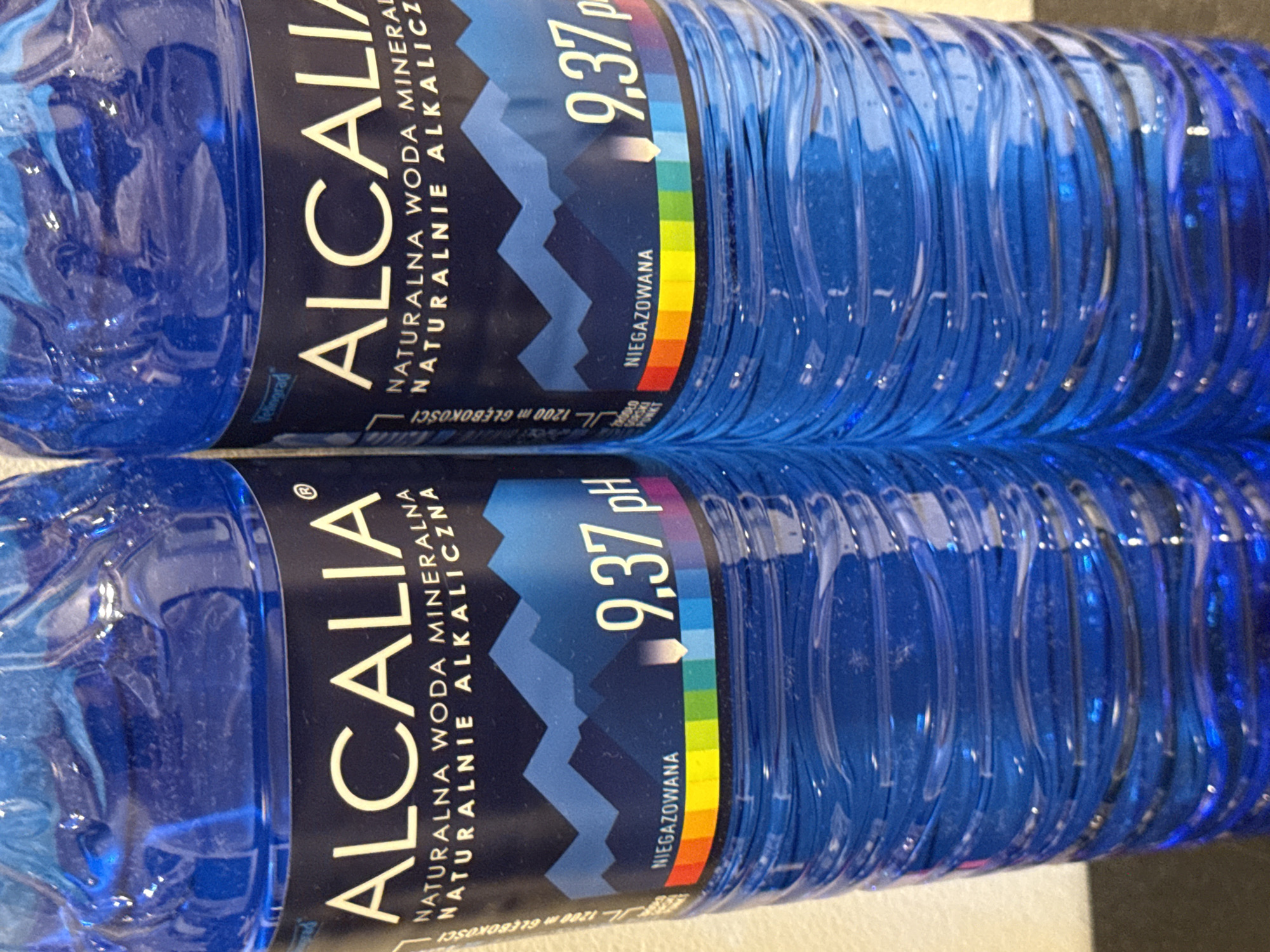 Two blue plastic bottles of ALCALIA natural mineral water are shown, standing next to each other. The bottles have dark blue labels with white text. The text on the label includes "ALCALIA", "NATURALNA WODA MINERALNA", "NATURALNIE ALKALICZNA", and "NIEGAZOWANA". The bottles display a mountain graphic and a pH reading of 9.37. A rainbow-colored bar is also visible on the label, indicating the pH level. The bottles appear to be in a bright, possibly indoor setting.