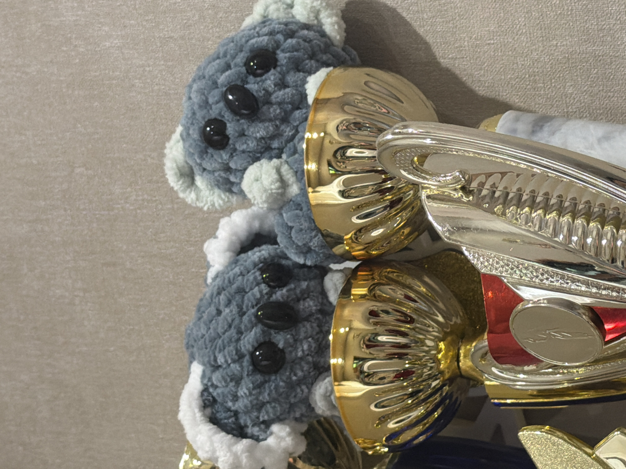 The image shows a close-up of two crocheted koala figures with black button eyes, sitting atop a stack of gold trophies. The koalas are gray with white and light green accents. The trophies are shiny and gold-colored, with intricate detailing. The background is a plain, textured wall. The scene is likely indoors, possibly a home or a display cabinet, given the presence of trophies and decorative items. There is no visible indication of the city of Dnipro, Ukraine, or any other specific geographic location.